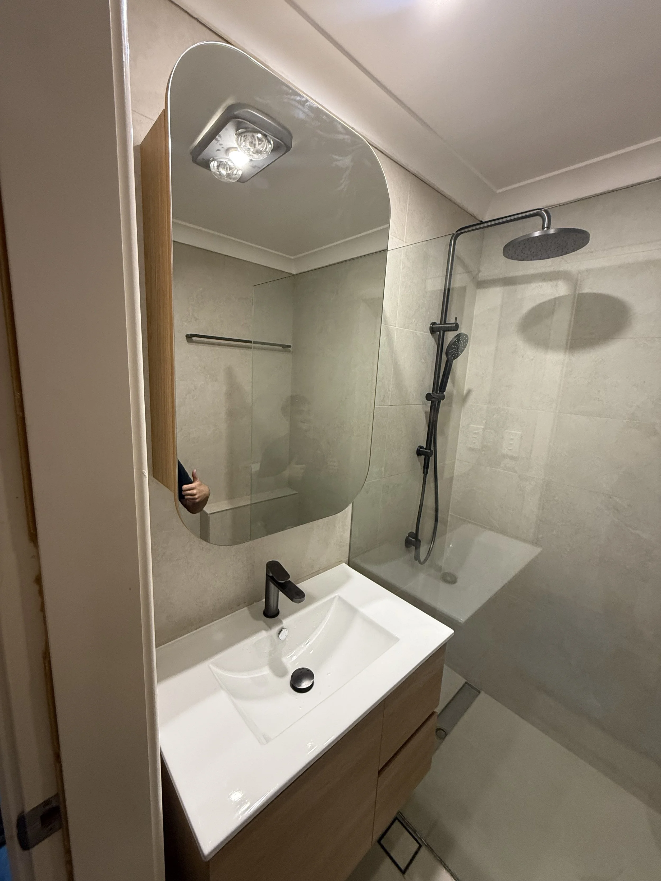 Bathroom with a white sink, a wooden cabinet, a rectangular mirror, a black faucet, and a gray shower with a rain showerhead and a handheld showerhead. The bathroom has beige tiled walls and white ceiling.