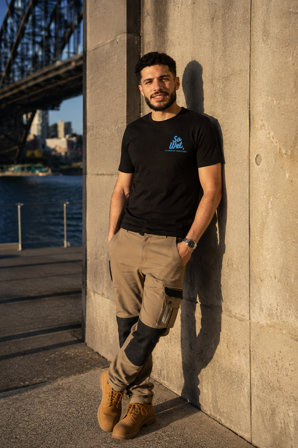 A man leaning against a concrete wall near a body of water, wearing a black t-shirt, beige cargo pants, and tan boots, with a bridge visible in the background.