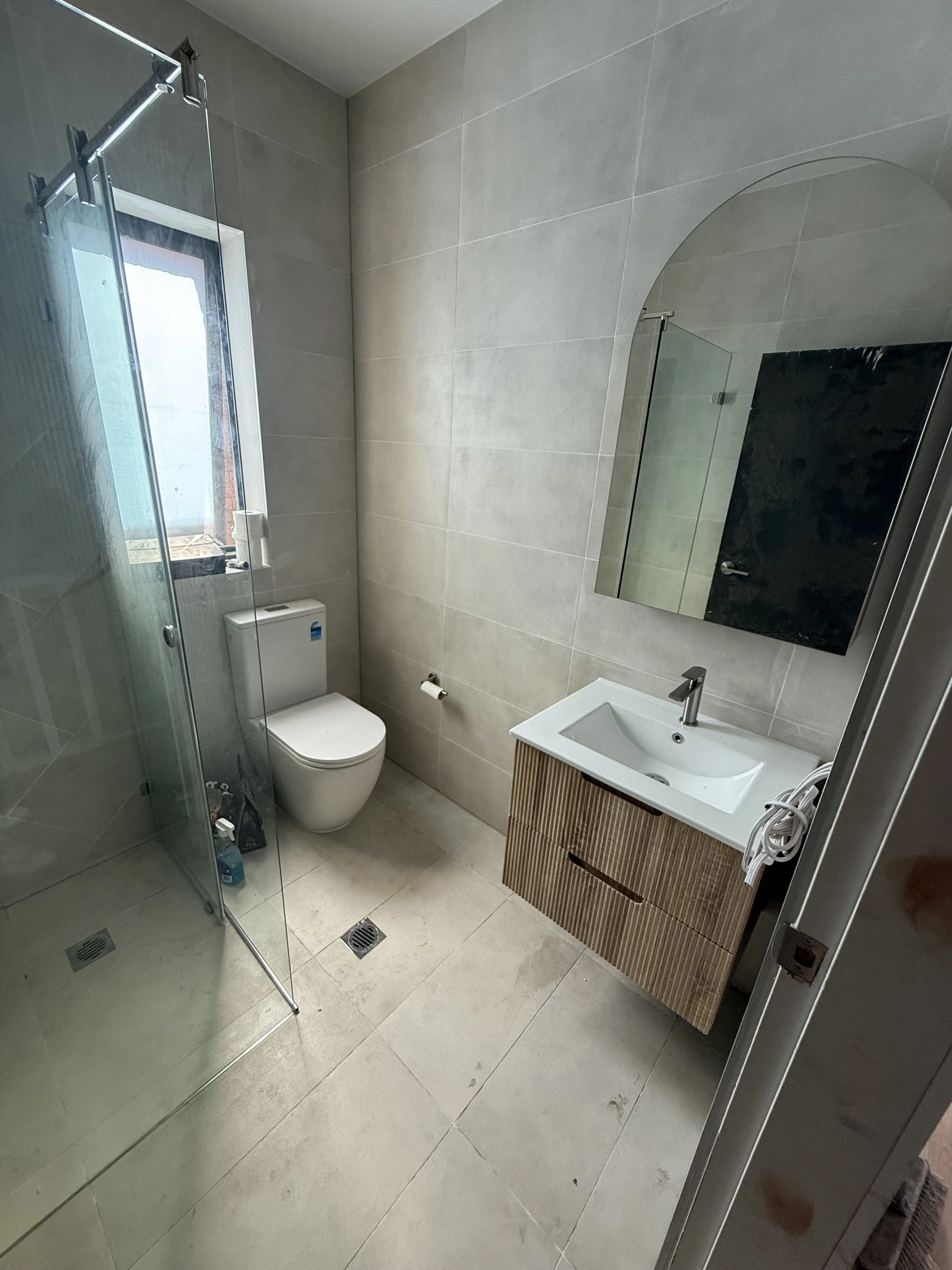 Modern bathroom with a shower enclosure, toilet, small sink with a wooden cabinet, a mirror, and beige tiled walls and floor.
