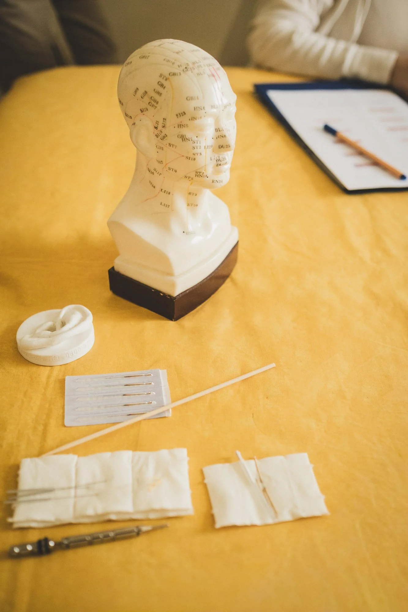Ein Kopfmodel aus Kunststoff mit Akupunkturmarkierungen, auf einem Tisch mit medizinischen Materialien wie Pflastern, Nadeln, Zungenabdruck, und einem Stift.