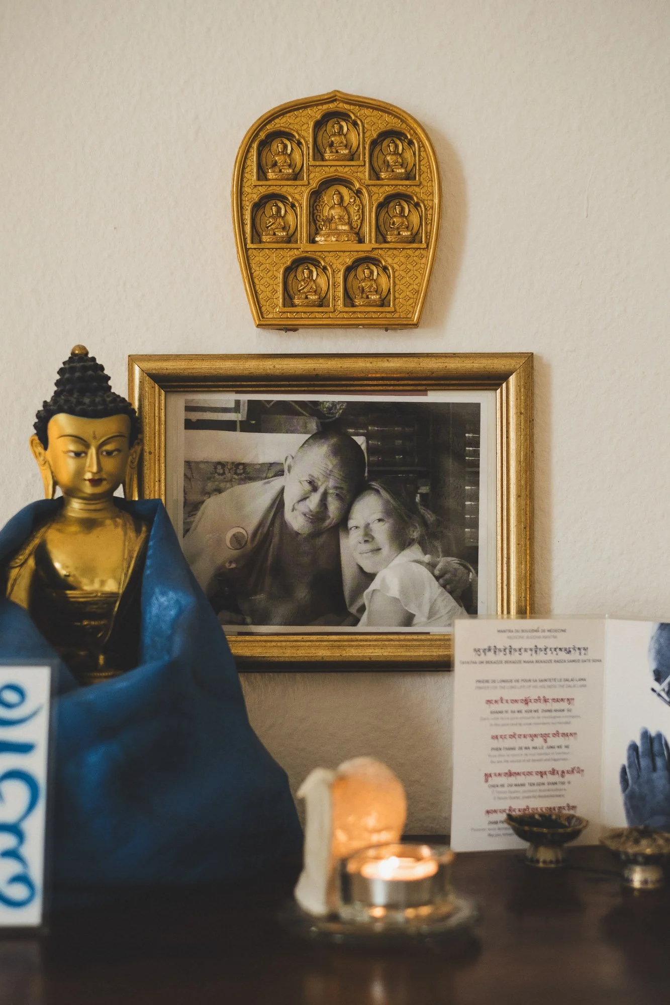 Ein Buddha-Statue in goldener Kleidung im Vordergrund, im Hintergrund ein schwarz-weißes Foto eines Mönchs und einer Frau, an der Wand hängen kleine Buddha-Statuen in goldfarbenen Rahmen.