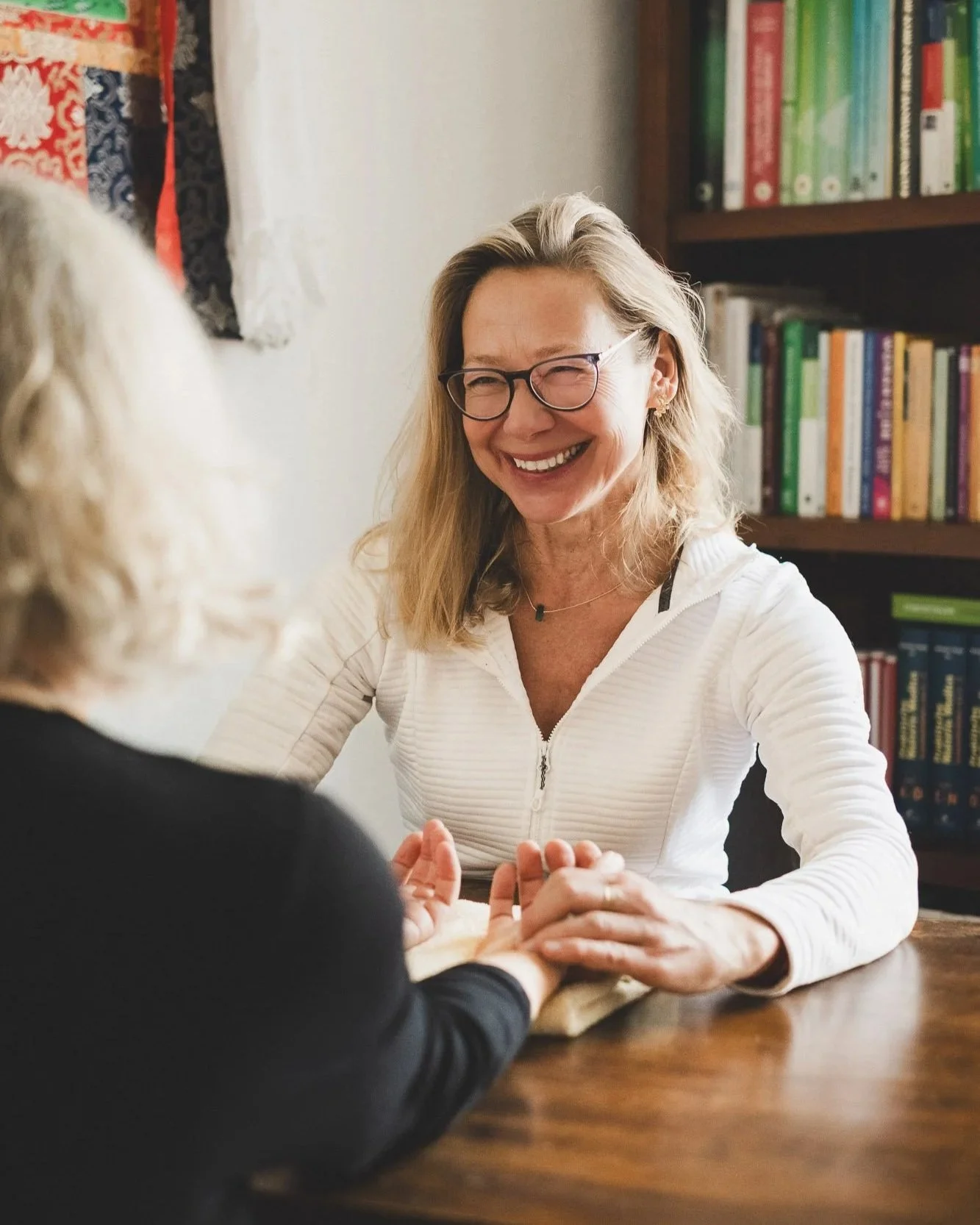 Zwei Frauen sitzen an einem Holztisch und haben ein Gespräch. Die Frau auf der rechten Seite hat blonde Haare, trägt eine weiße Jacke und trägt Brille. Im Hintergrund sind Bücherregale mit bunten Büchern.