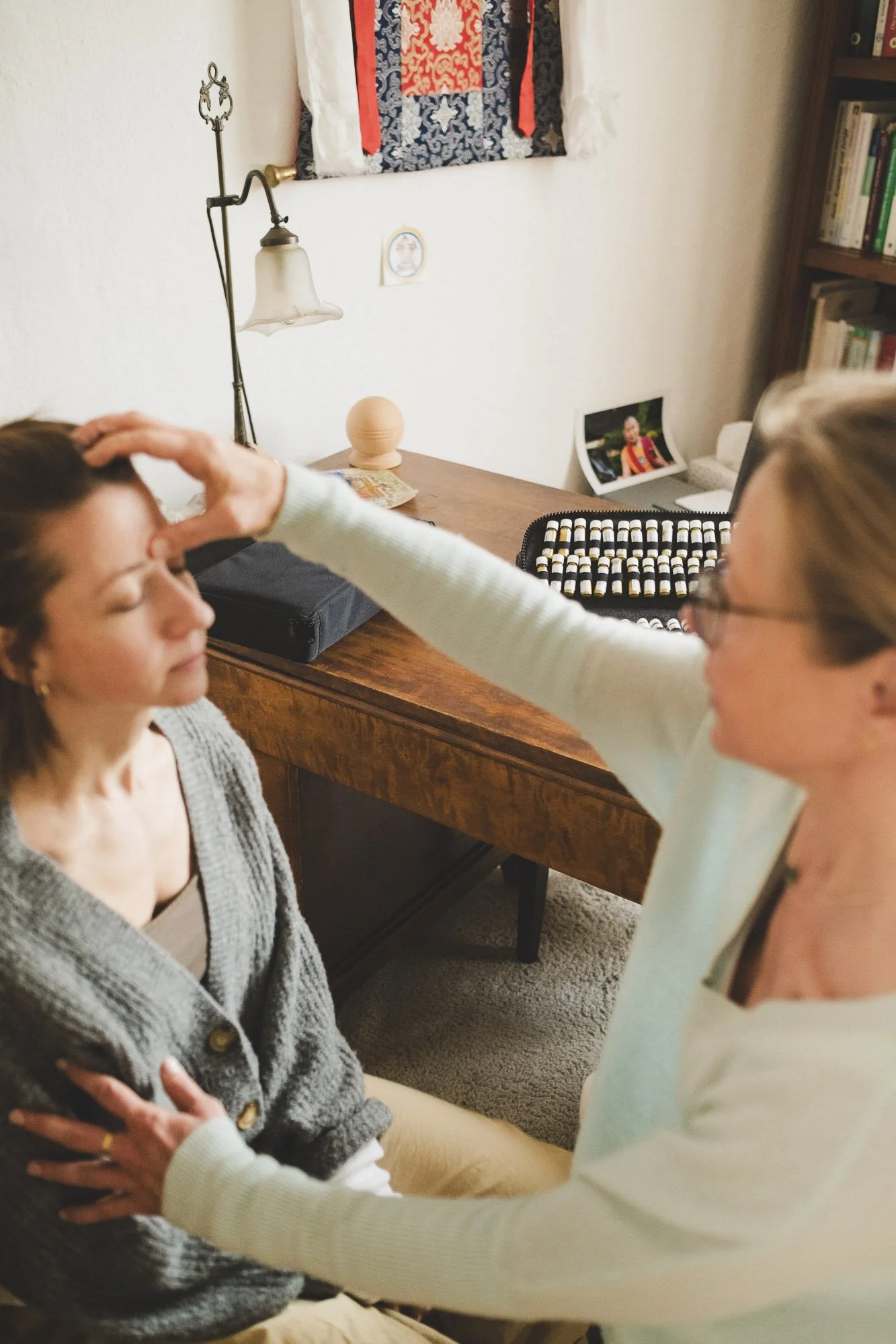 Zwei Frauen sitzen sich gegenüber in einer Praxis. Eine Frau hat braunes Haar und trägt einen grauen Pullover. Die andere Frau trägt eine Brille und einen weißen Pullover, diese berührt die andere Frau auf der Stirn.