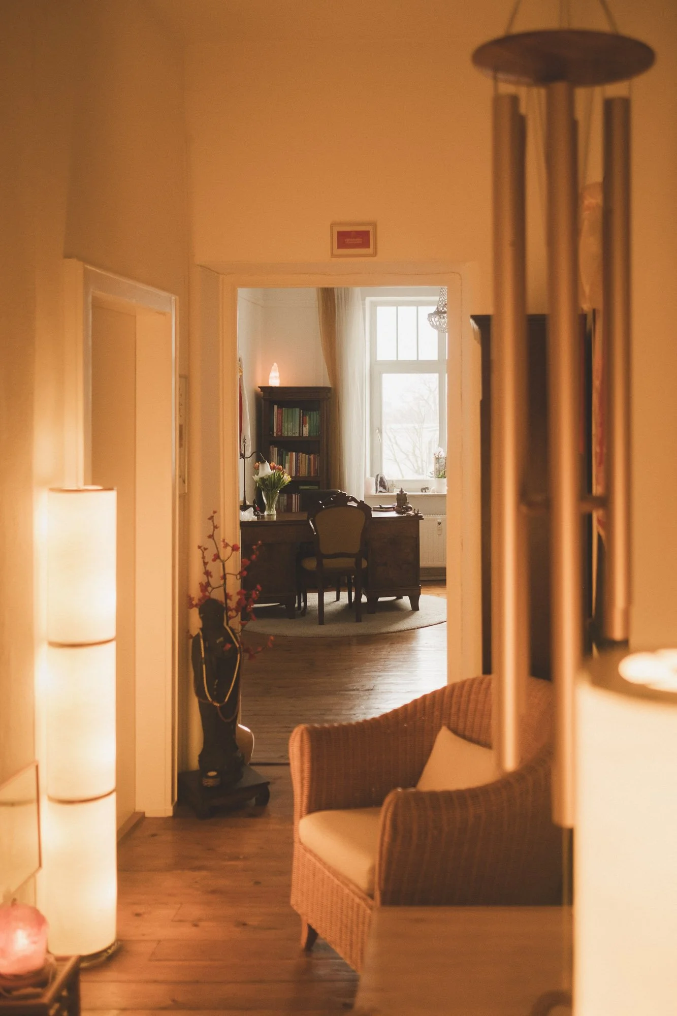 Einblick in ein gemütliche  mit Holzfußboden, Wänden und Möbeln im klassischen Stil. Im Hintergrund ein Schreibtisch mit Stuhl, Regal mit Büchern, eine Vase mit Blumen und große Fenster mit Vorhängen. Im Vordergrund ein braunes Armchair, eine Stehlampe und ein kleines Beistelltischchen mit einem Salzlampen-Objekt.