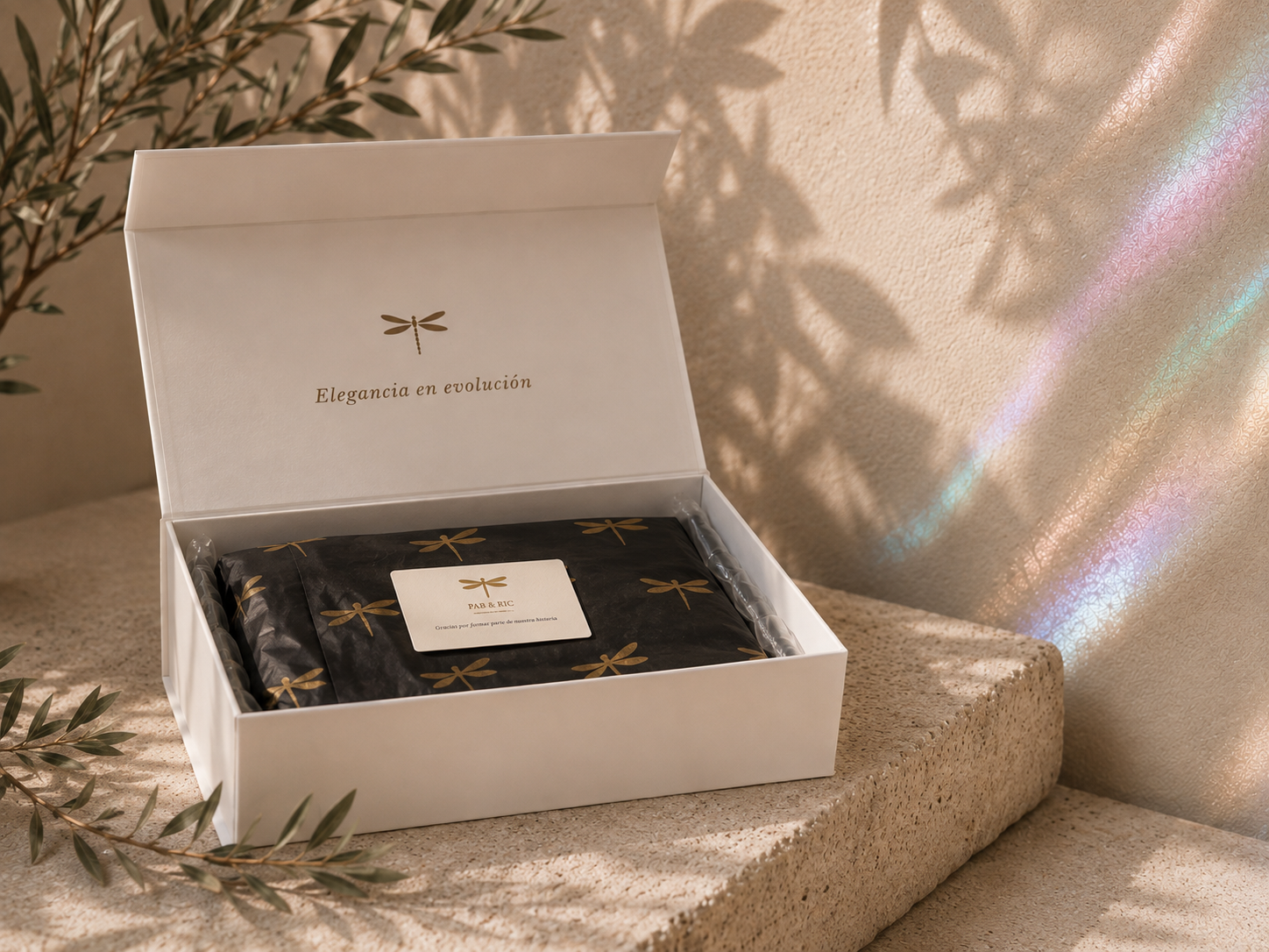 Una caja blanca abierta con interior decorado con un diseño de libélulas doradas y la frase "Elegancia en evolución" en la tapa. Dentro, hay un paquete envuelto en papel negro con libélulas doradas, y una tarjeta con el logo y la frase "Gracias por formar parte de nuestra historia." La caja está sobre una superficie de piedra, con ramas verdes a un lado y sombras de hojas proyectadas en la pared de fondo, que refleja luz y crea un arcoíris suave.