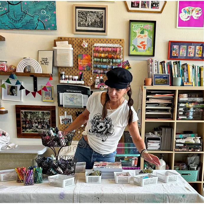 A woman in a black hat and t-shirt with a floral print, standing at a table with baskets and containers of pens, markers, and craft supplies in an art studio or craft room.