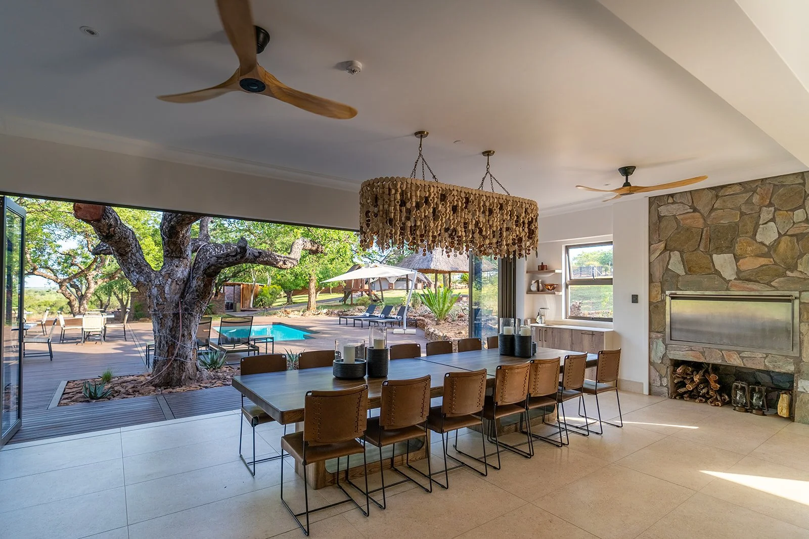 Indoor dining room with a large wooden table, eight chairs, a stone fireplace, and a large open window showing a backyard with a pool, patio furniture, trees, and greenery.