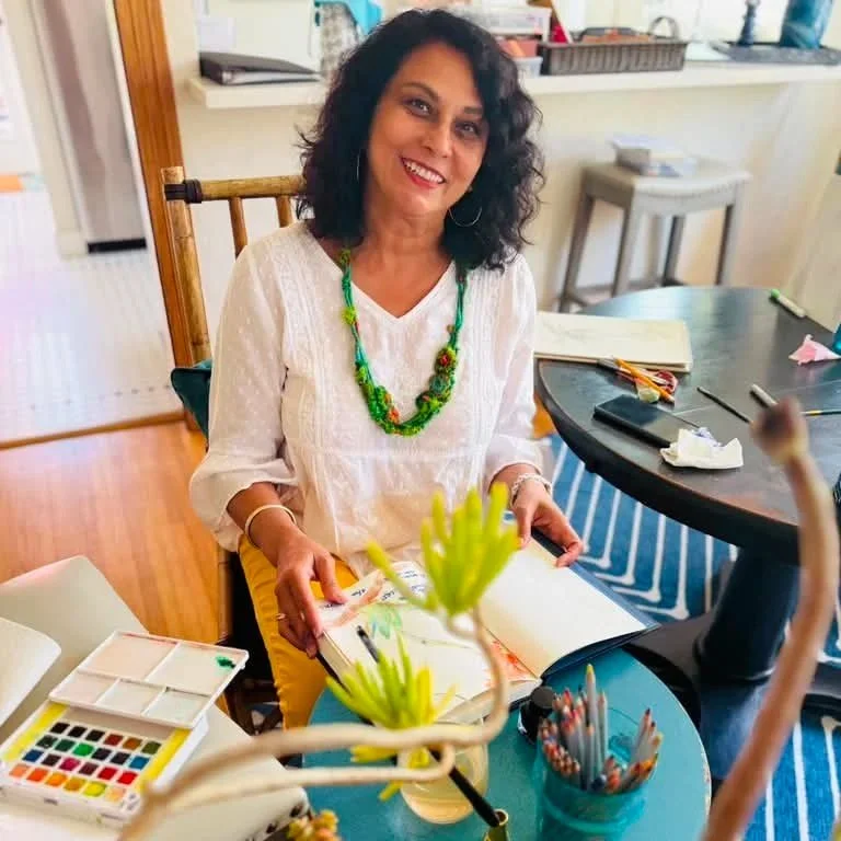 A woman with curly black hair wearing a white top and colorful necklace sitting at a table with art supplies, smiling at the camera.
