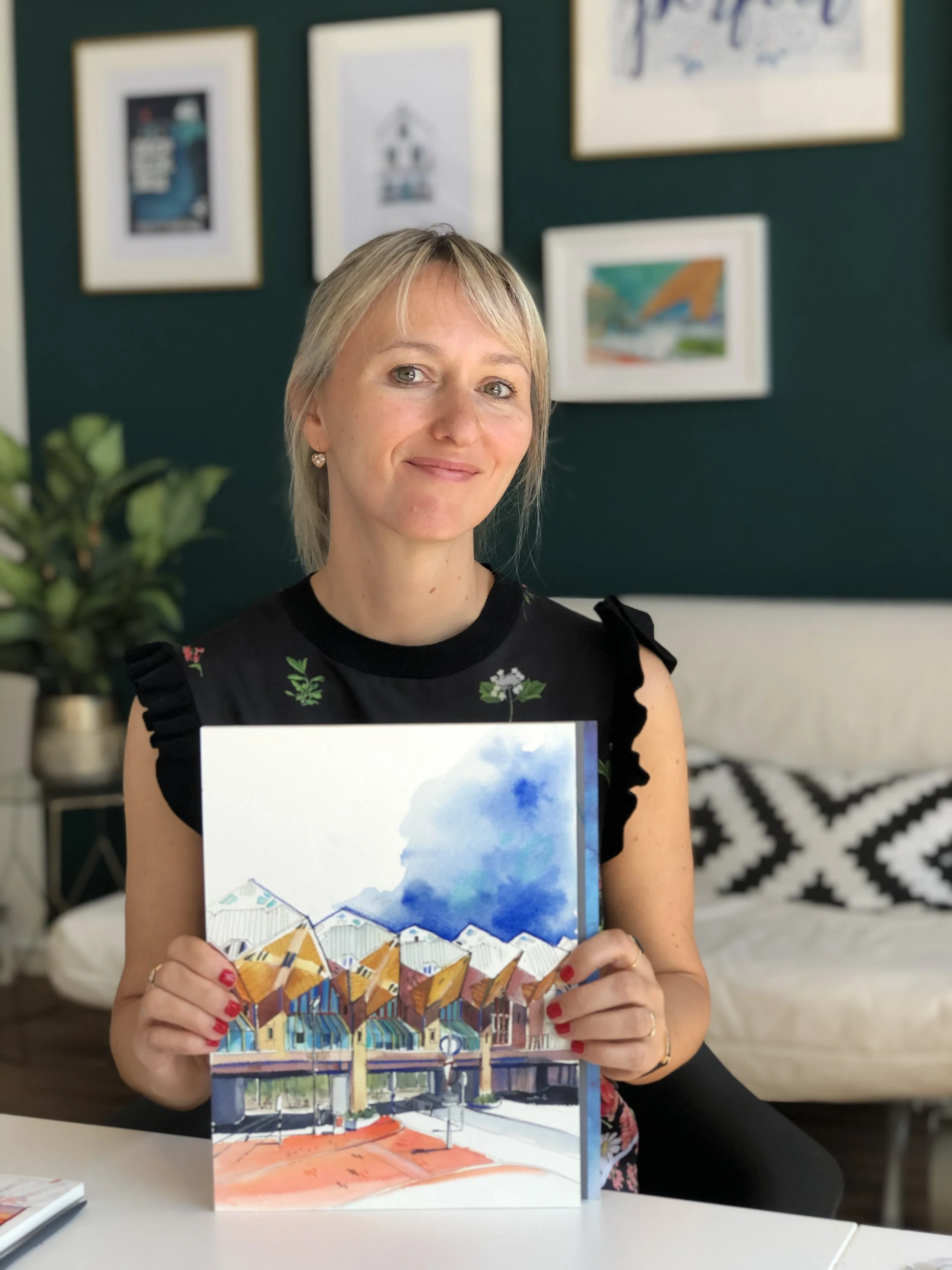 Woman with blonde hair holding a colorful painting of a building with bright yellow, orange, and blue rooftops outdoors.