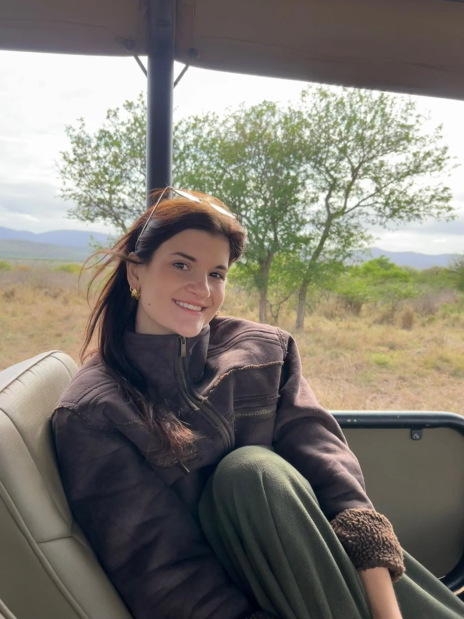 A woman with long brown hair, wearing a brown jacket and earrings, sitting in a vehicle with a window showing a landscape of trees and mountains behind her, smiling at the camera.