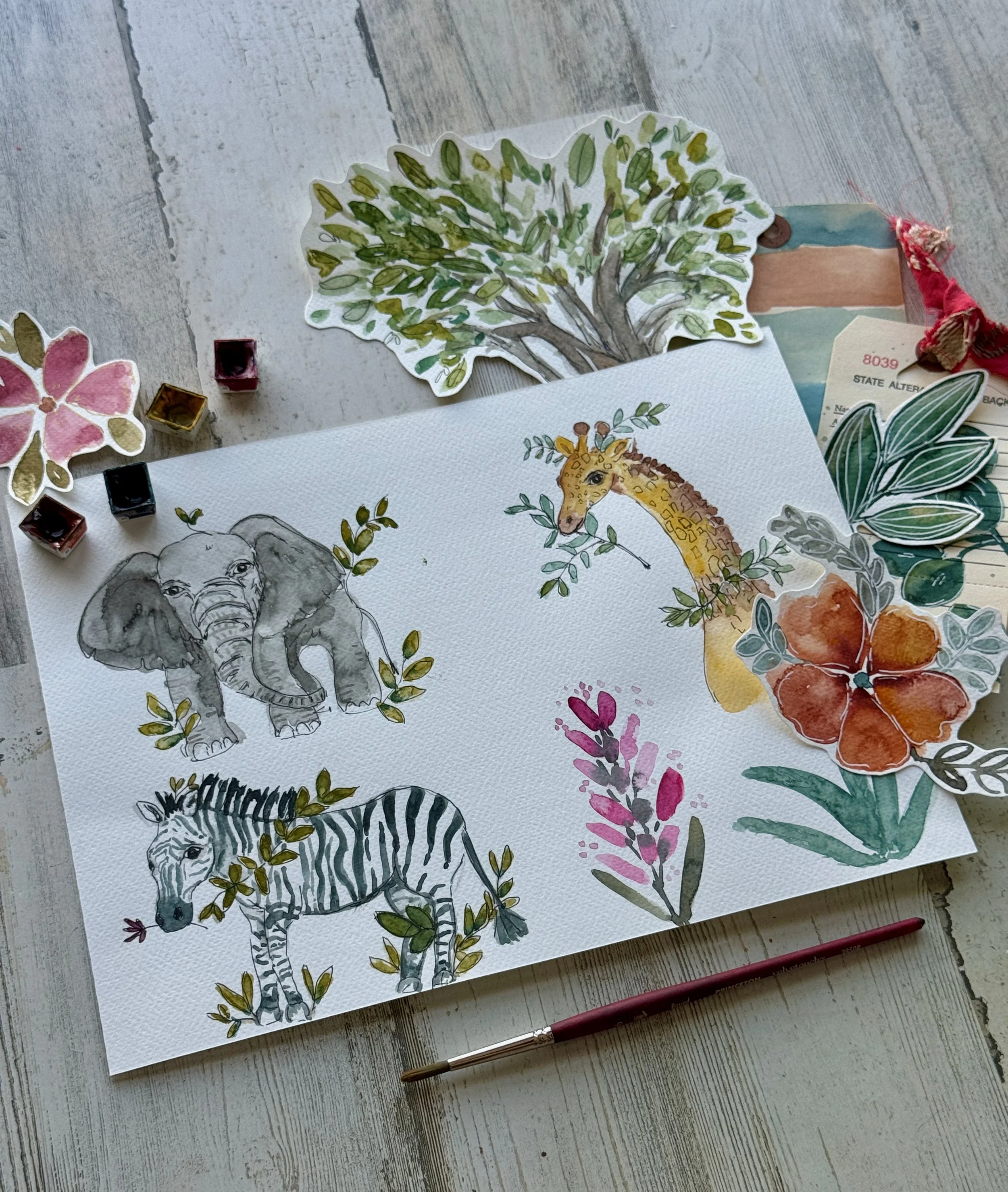 Watercolor paintings of African animals including an elephant, a giraffe, and a zebra, surrounded by leaves and floral patterns, placed on a wooden table with watercolor brushes nearby.