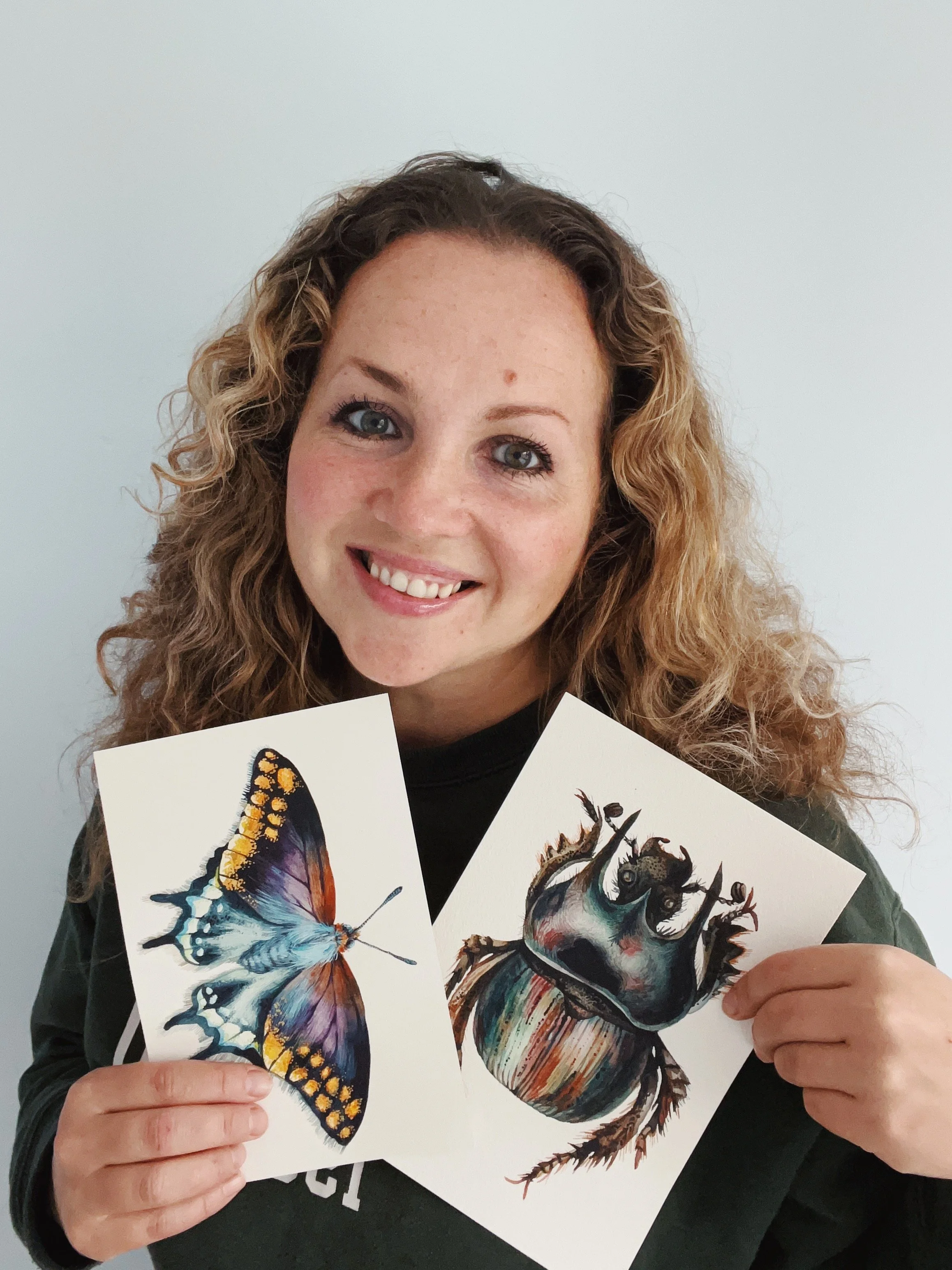 Person with curly hair smiling while holding illustrations of a butterfly and a beetle.