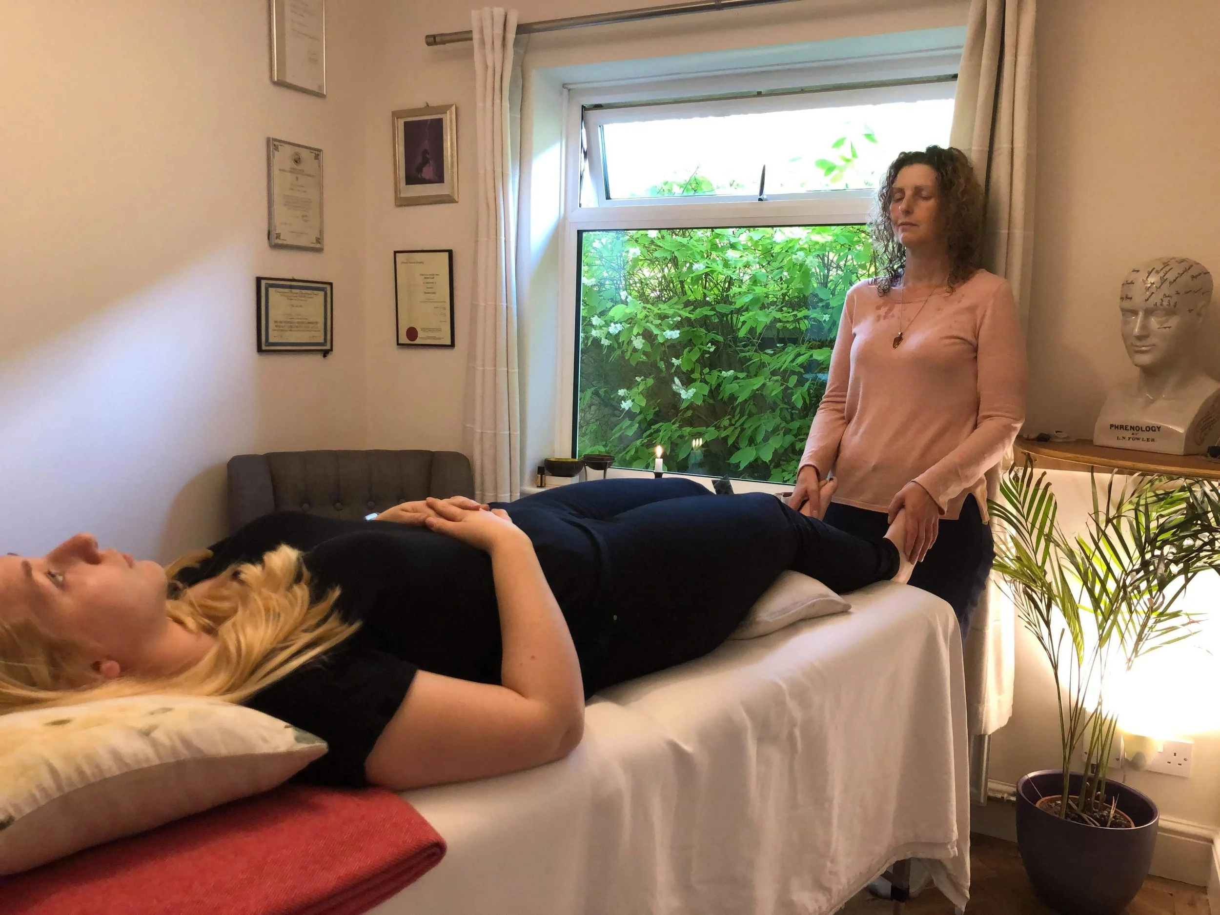A woman (therapist, Jayne) is standing at the end of the couch, holding the feet of another woman (patient lying on a couch, with her eyes closed and her hands resting on her abdomen. Several framed certificates on wall of comfortable treatment room