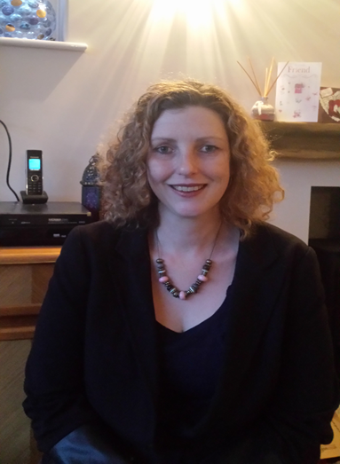 A woman with curly red hair smiling, wearing a dark top and a beaded necklace, sitting indoors with a fireplace, decorative items, and a shelf in the background. Therapist