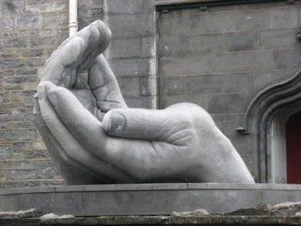 A large stone sculpture of a hand positioned with fingers folded and thumb extended, set against a brick building background.