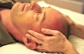 Close-up of a man receiving a specific craniosacral hold, with therapists hands at the sides of the man's head and fingers resting gently forward and behind ears. Man's eyes closed, relaxed expression.