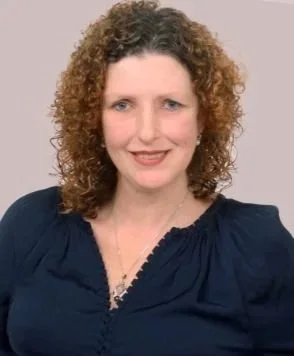 Portrait of a woman with curly, shoulder-length hair wearing a dark blue blouse and a necklace, smiling against a plain background. Jayne the Craniosacral Therapist and Reiki Healer