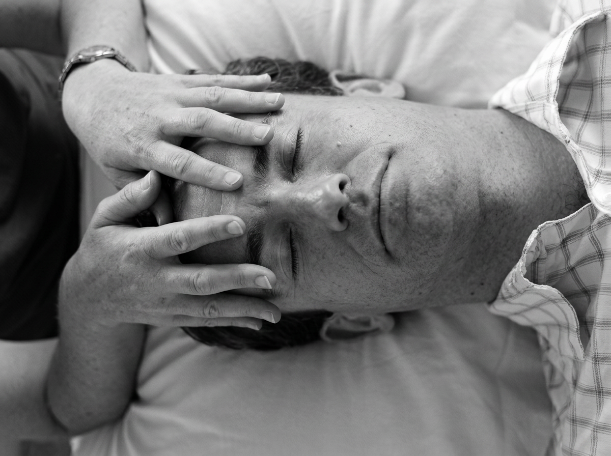 A person lying on their back with eyes closed, receiving a massage or treatment on their forehead from a professional with hands gently placed on their head.