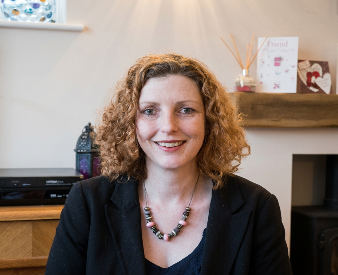 A woman with curly red hair smiling, wearing a black blazer and a colorful beaded necklace, sitting indoors with a wooden shelf and greeting cards in the background. Jayne Craniosacral Therapist and Reiki Healer