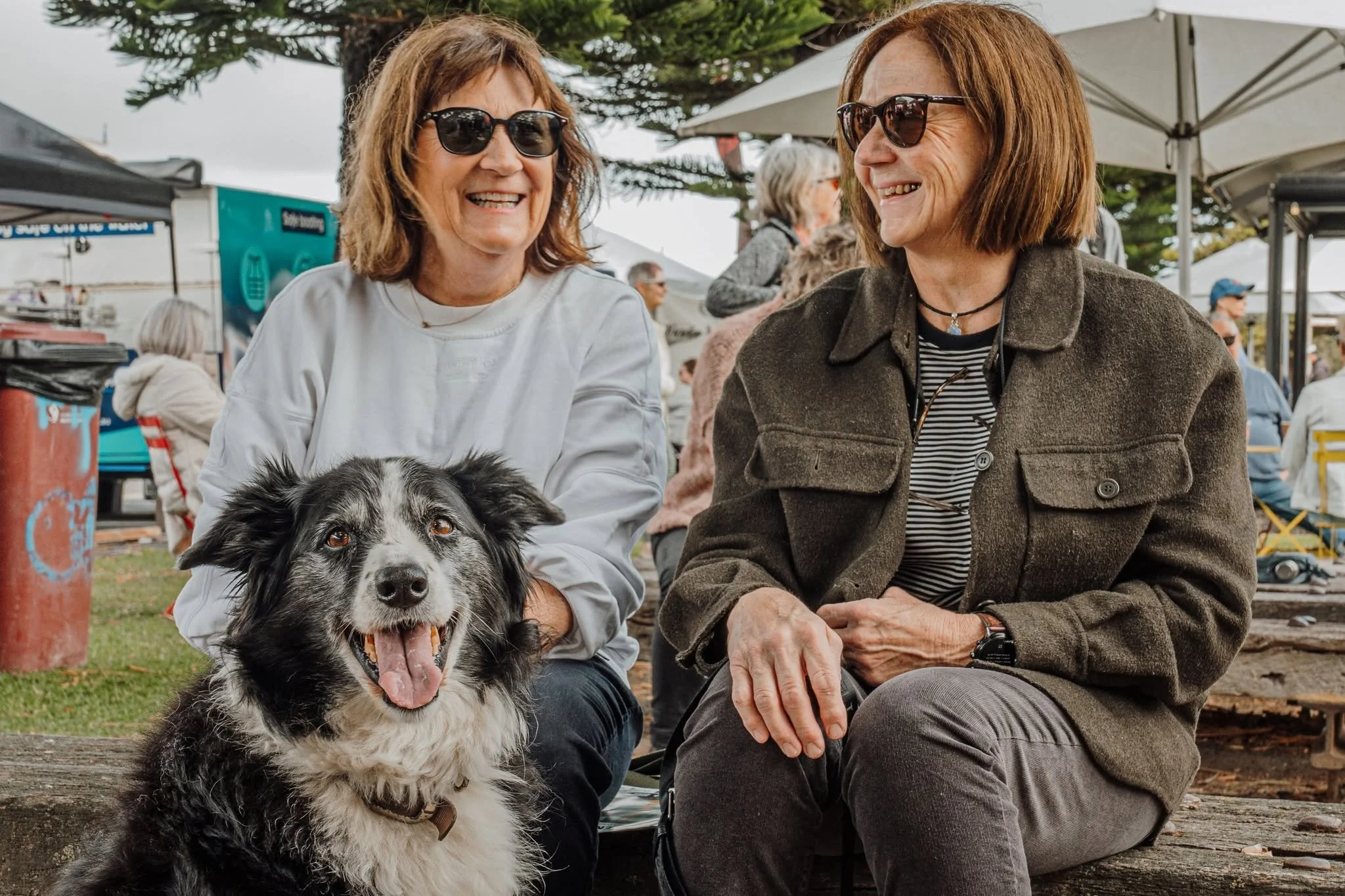 Two women wearing sunglasses sitting outdoors at the Festival, smiling at each other, with one holding a black and white dog that is yawning.