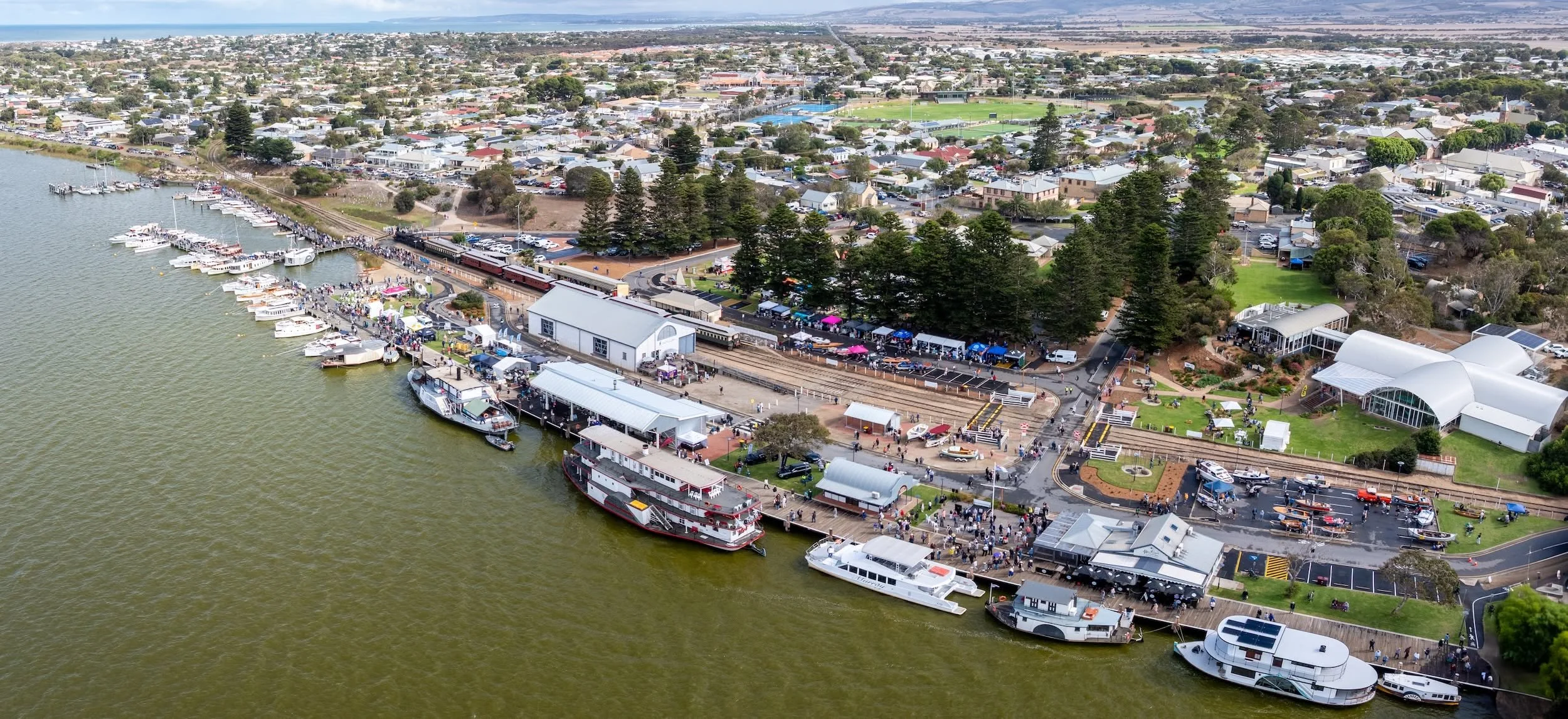 Aerial view of the Goolwa Wharf with boats docked, a crowded marketplace, and a train passing through a park with trees and open spaces.