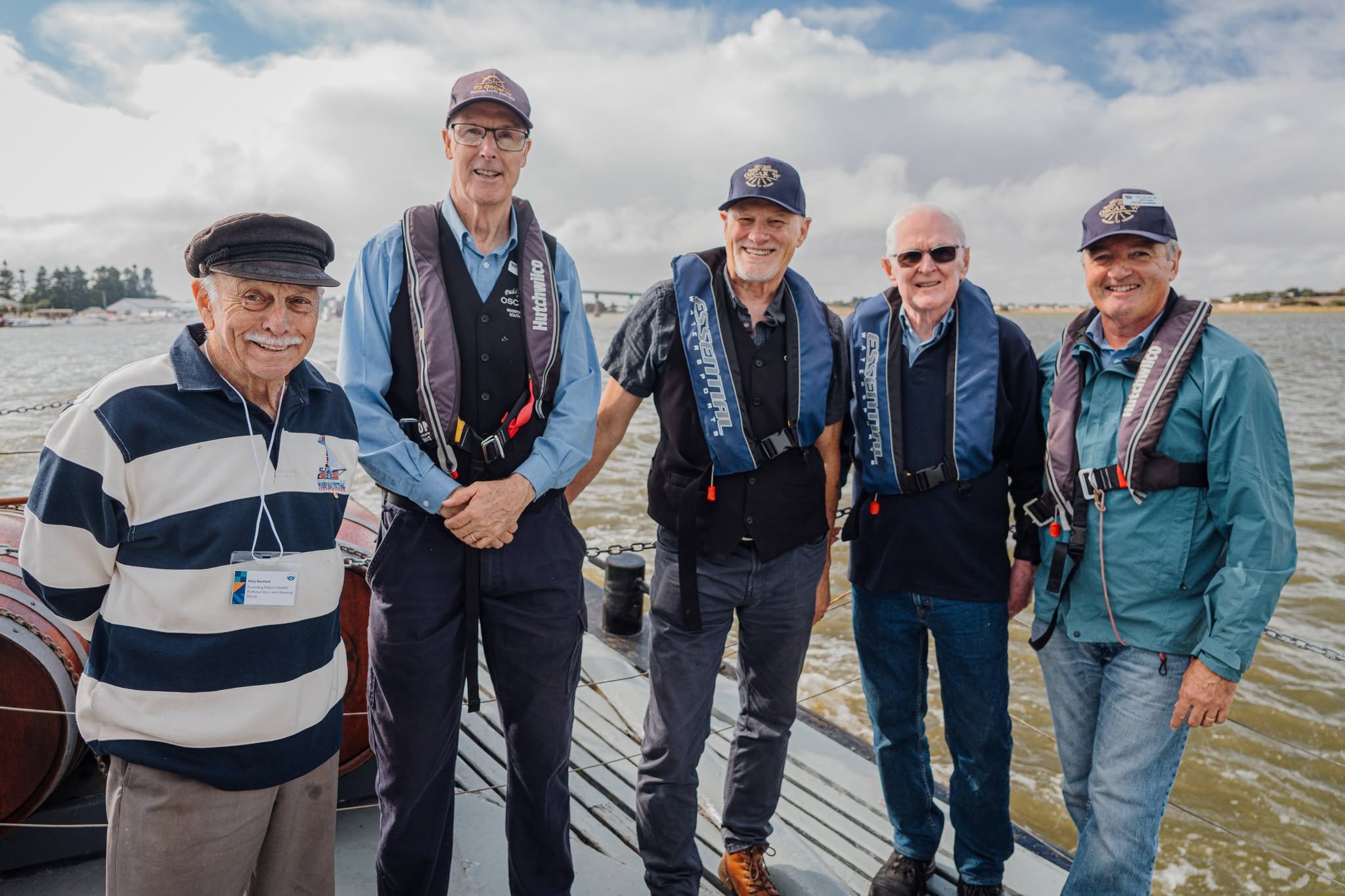 Five men standing together outdoors near water, smiling at the camera, with clouds and a bridge in the background.