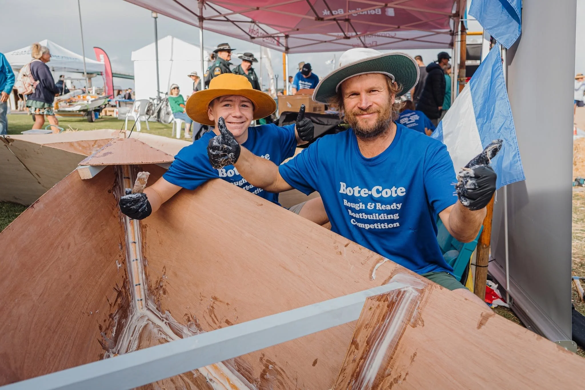 Two men wearing blue t-shirts and hats are sitting inside a wooden boat under a tent at an outdoor event. They are smiling and giving thumbs-up signs, with one man wearing gloves. Other people are walking and standing in the background.