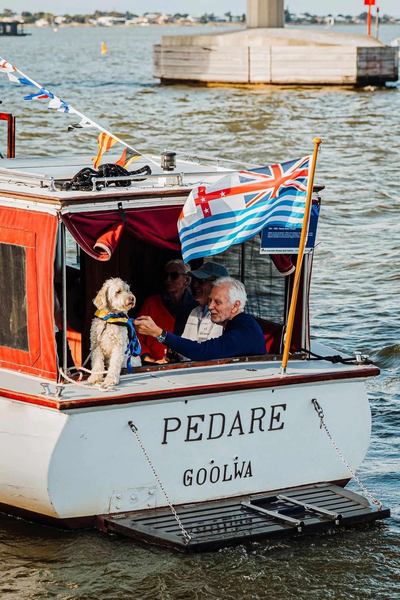 A boat named Pedare Goolwa decorated with Union Jack and Australian flags carries four people and a dog, sailing in a river with a bridge and distant shoreline visible in the background.