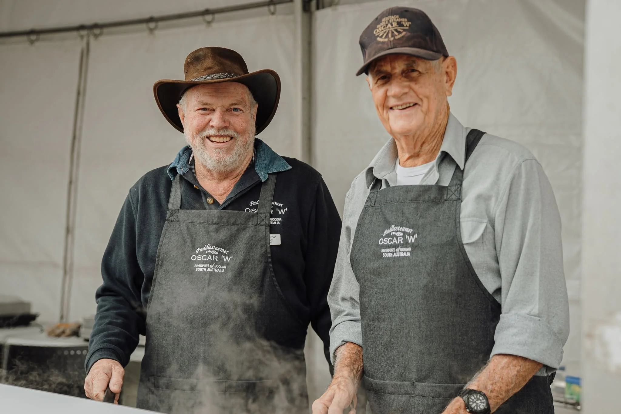 Two men wearing gray aprons, smiling and standing behind a food stall counter at the Festival
