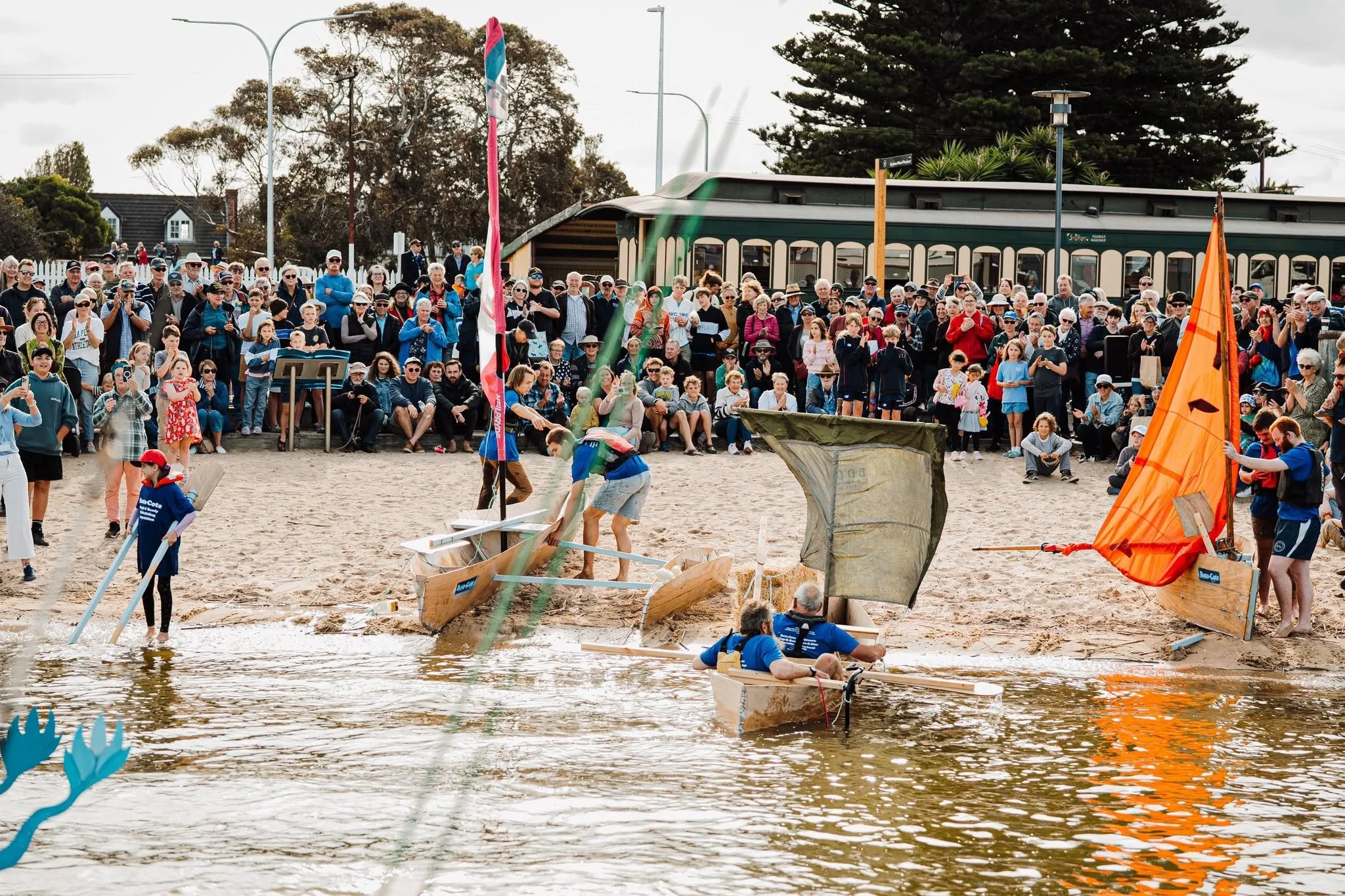 A large crowd of people gathered on a beach, watching a boat race involving small boats launching from the water onto the sand, with some boats visible in the water and wooden structures on the beach.