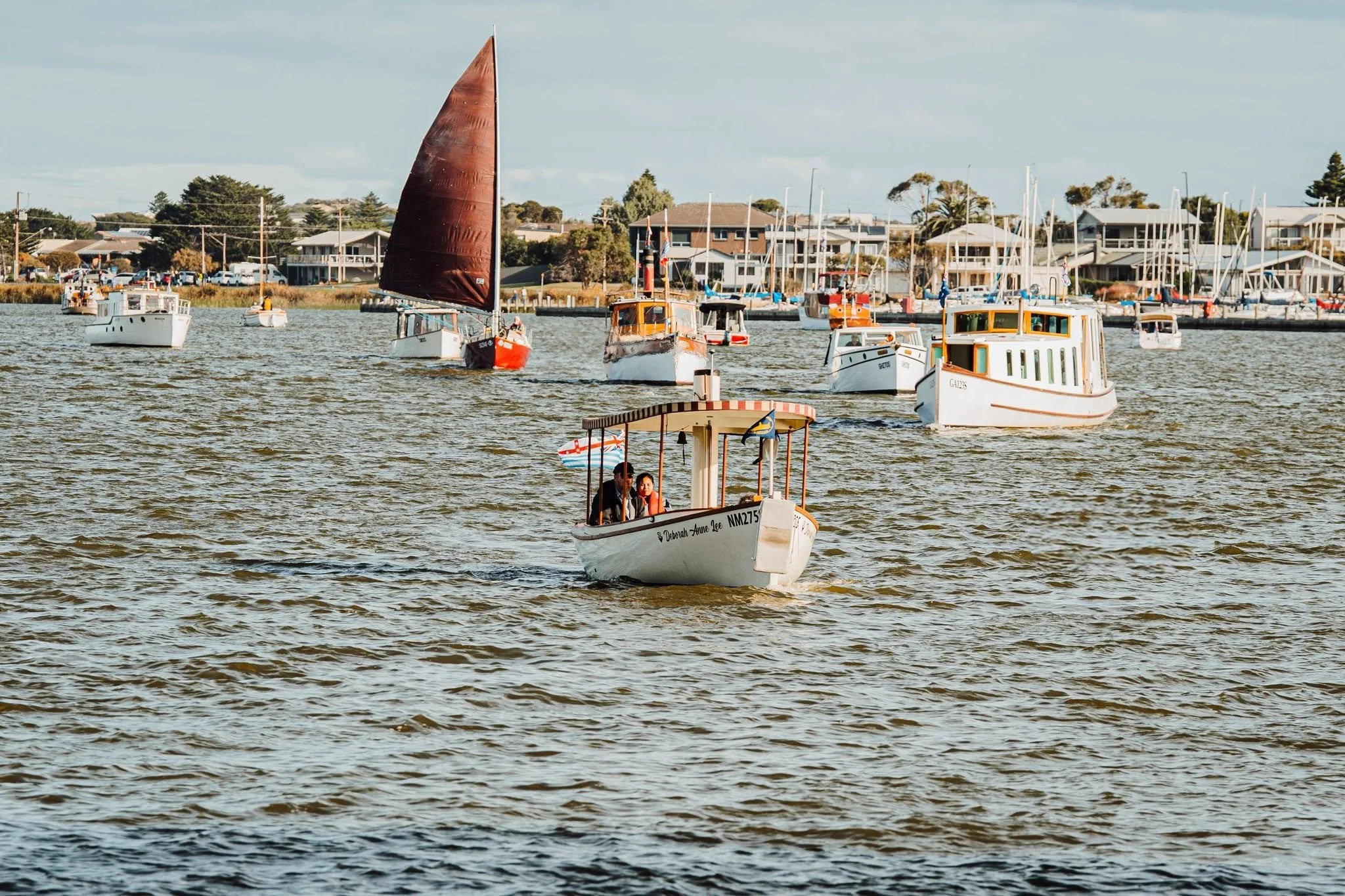 Several small boats sailing with houses and trees in the background.