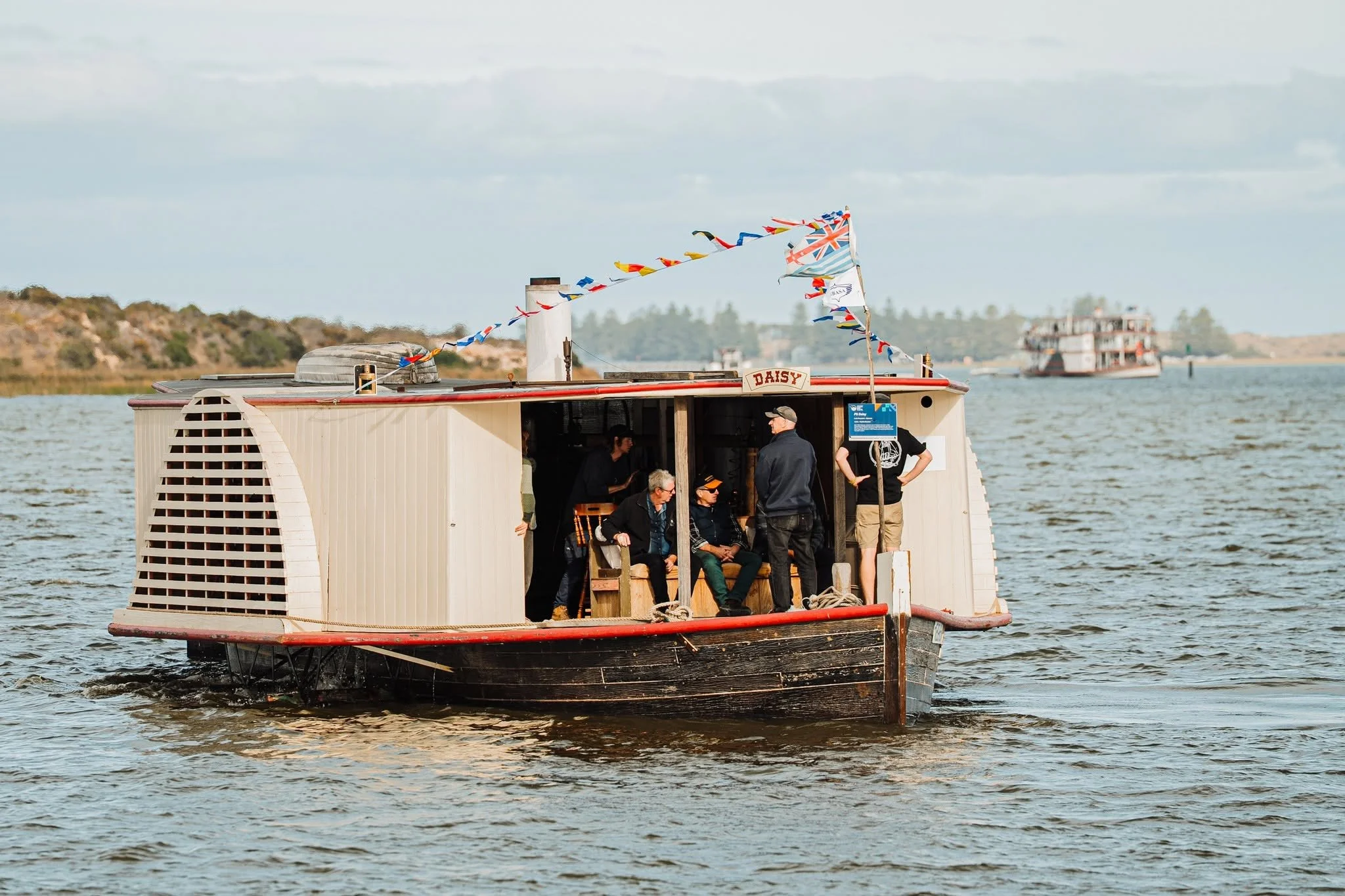 A boat named Daisy with people onboard, sailing during daytime. The boat is decorated with colourful flags and has a partially open cabin with visible people inside. In the background, there is a shoreline with trees and another boat in the distance.