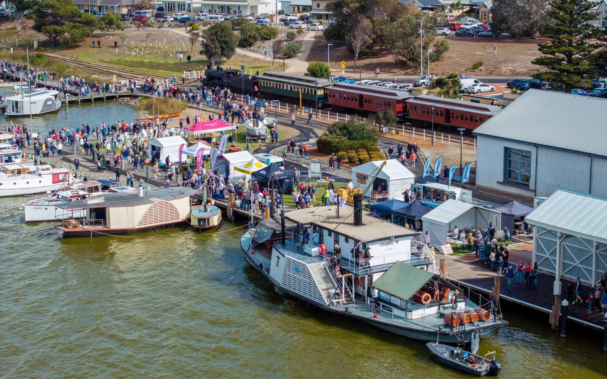A crowded Goolwa Wharf with people waiting in line to board the vintage steam train. There are boats docked along the water, tents, and vendor booths, with residential buildings and trees in the background.