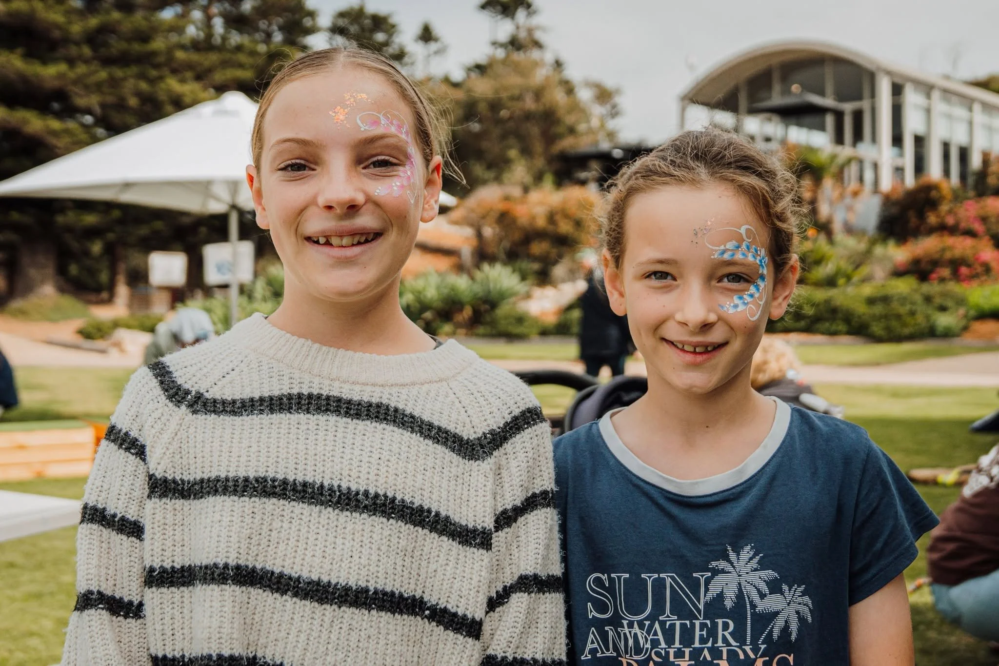 Two young girls with face paint standing outdoors at a park-like setting with trees, a building, and umbrellas in the background.