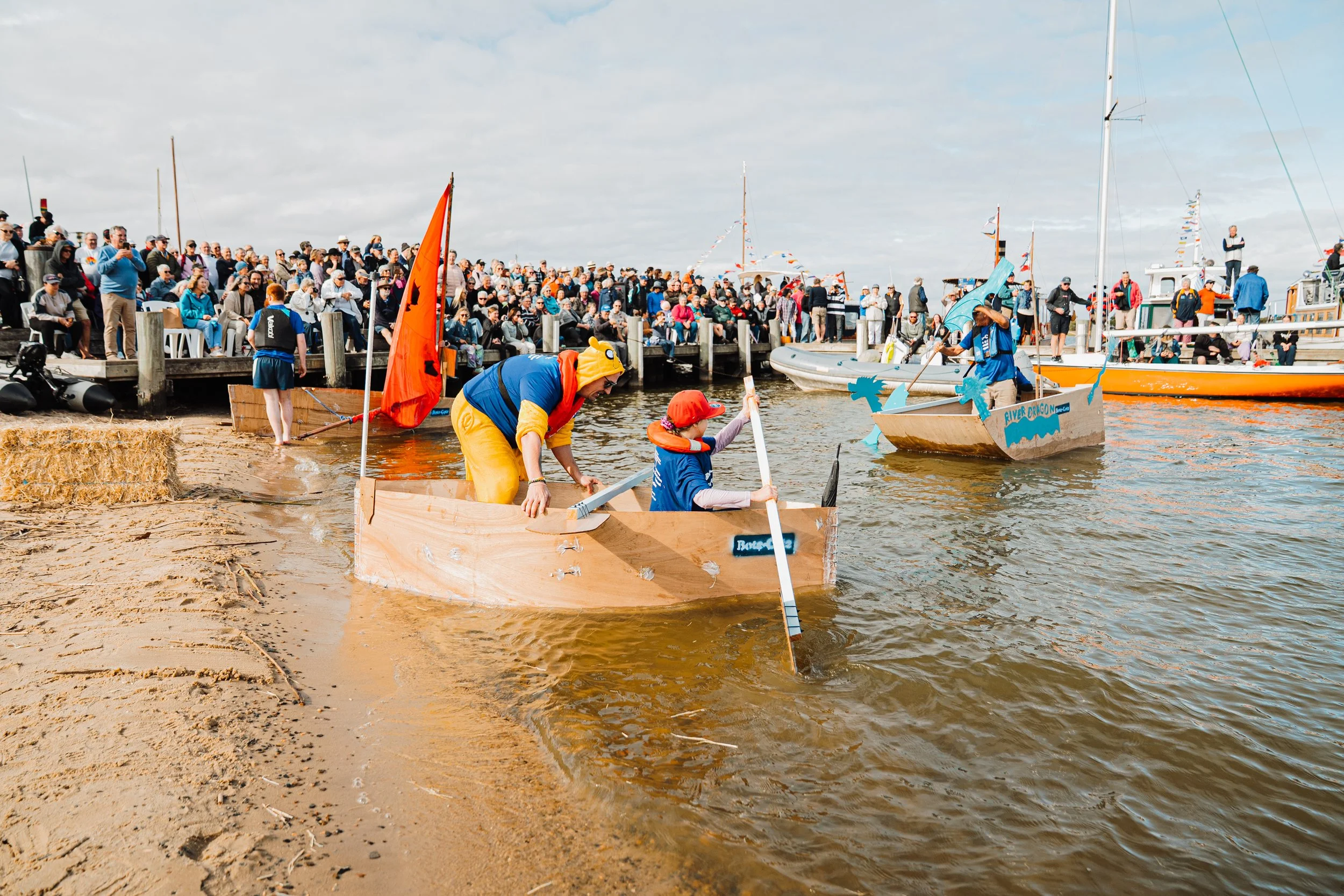 Crowd watching a boat race at a dock. Two children in cardboard boats are in the water, paddling with oars. The scene is outdoors with a cloudy sky.