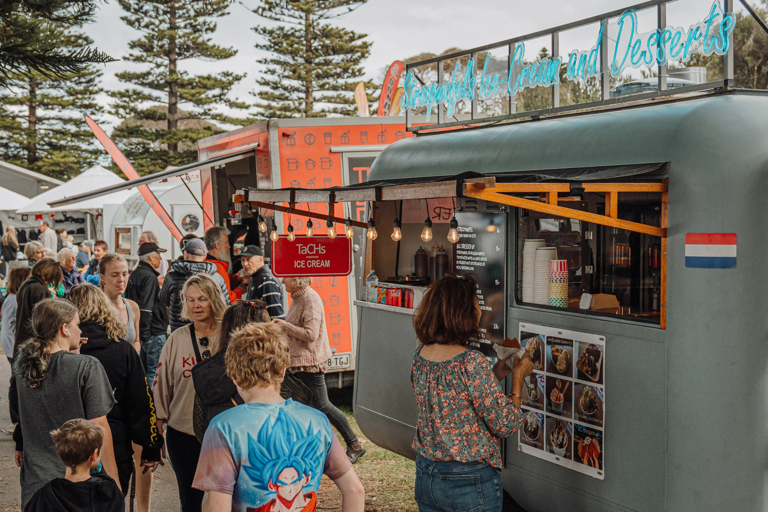 A crowd of people gathered around a food truck offering bubble tea, ice cream, and desserts at an outdoor event with trees in the background.