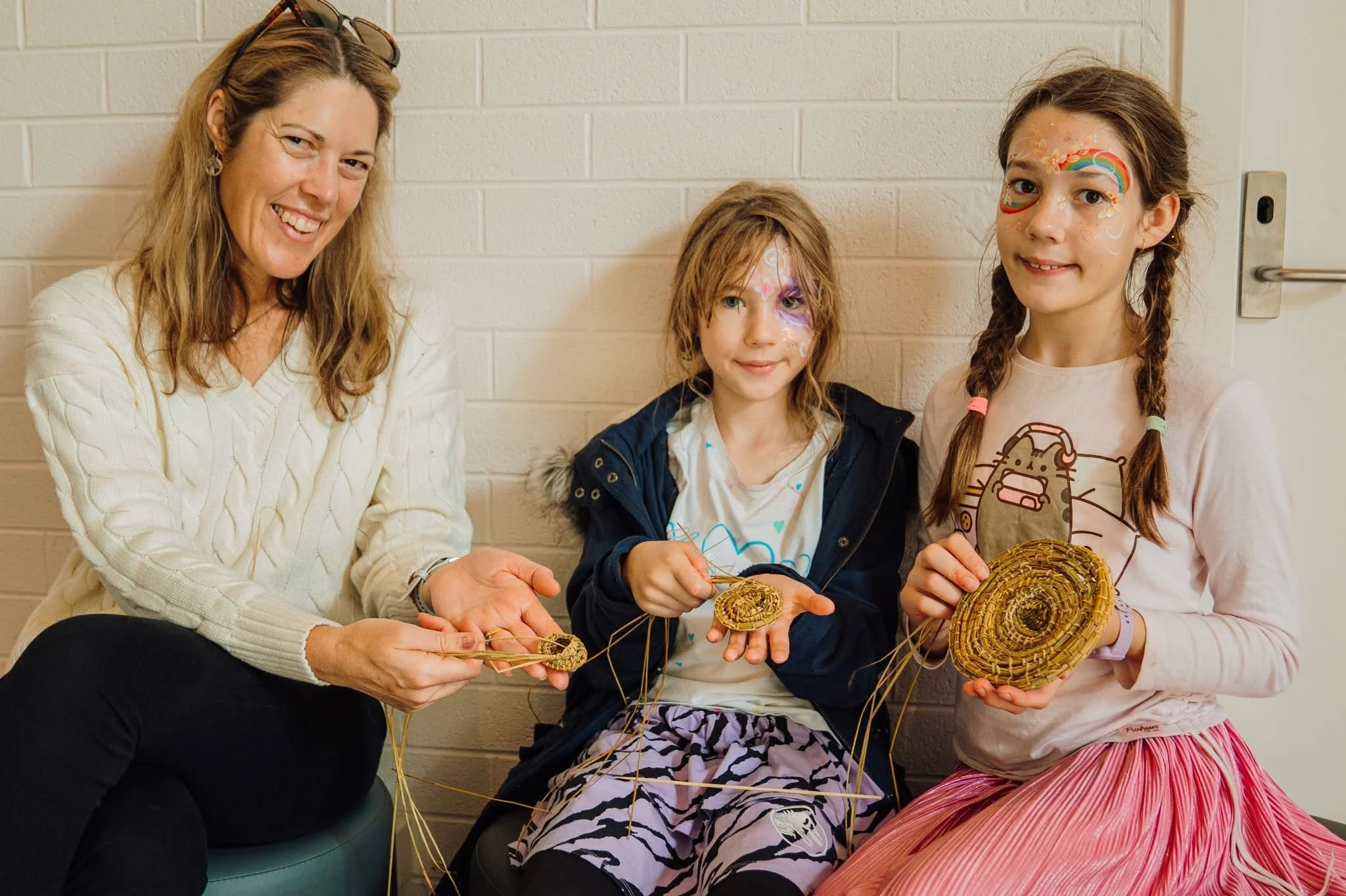 Three children and one woman sitting against a beige brick wall, making wreaths with straw and decorated with face paint and casual clothing.