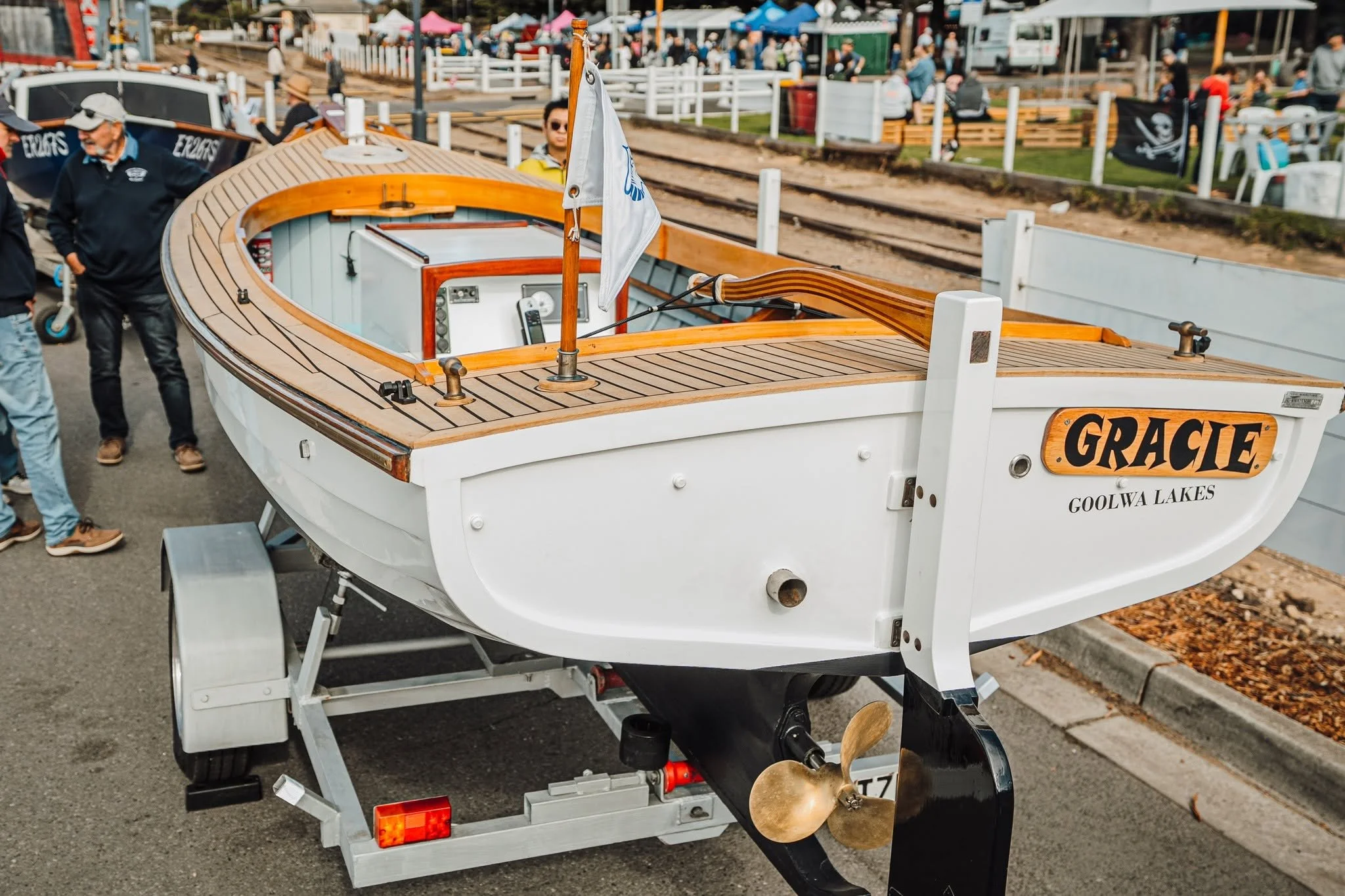 A wooden boat named 'Gracie' on a trailer at the Festival with people, tents, and railway tracks in the background.