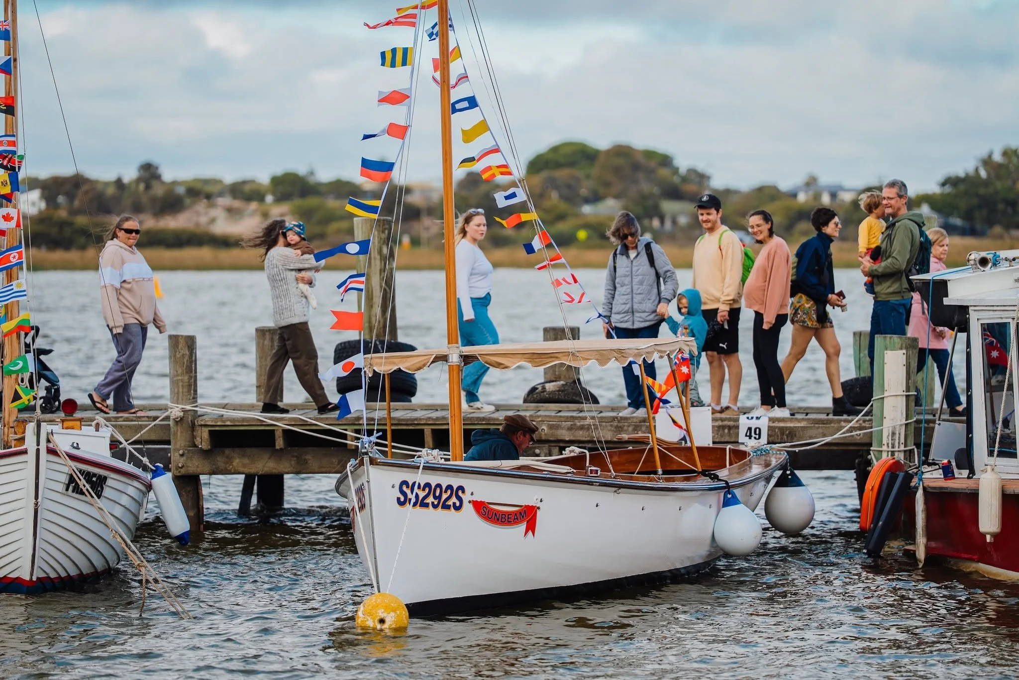 A group of people walking on a wooden dock by the water, with sailboats docked nearby decorated with colorful flags and maritime-themed banners, under a partly cloudy sky.