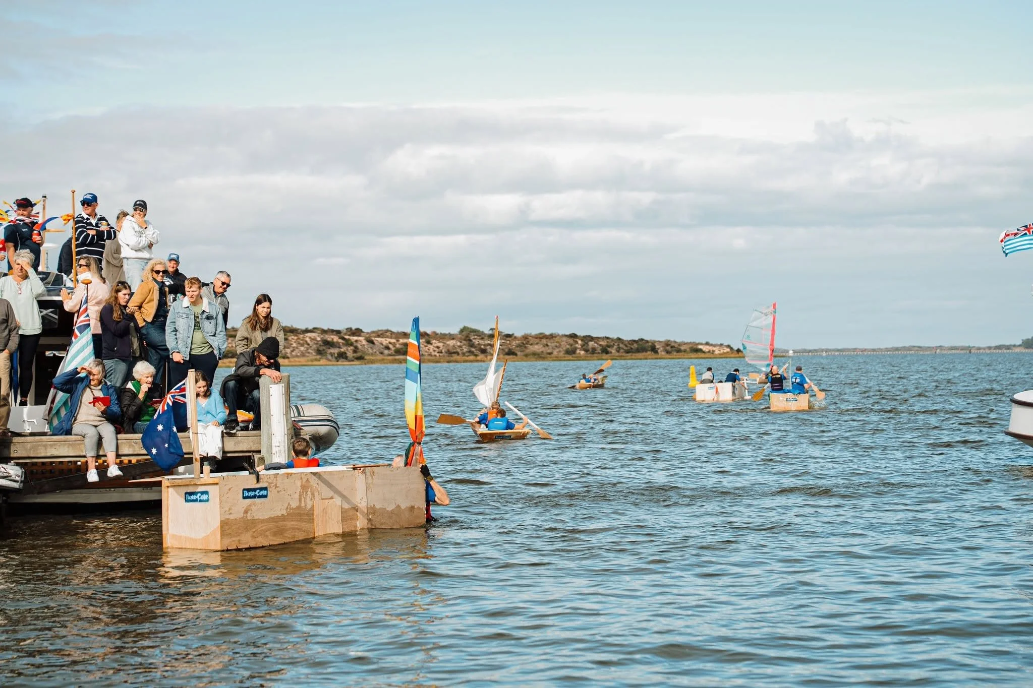 People standing on a dock watching sailboats, some children in small boats sailing on the water, and a windsurfer in the distance.