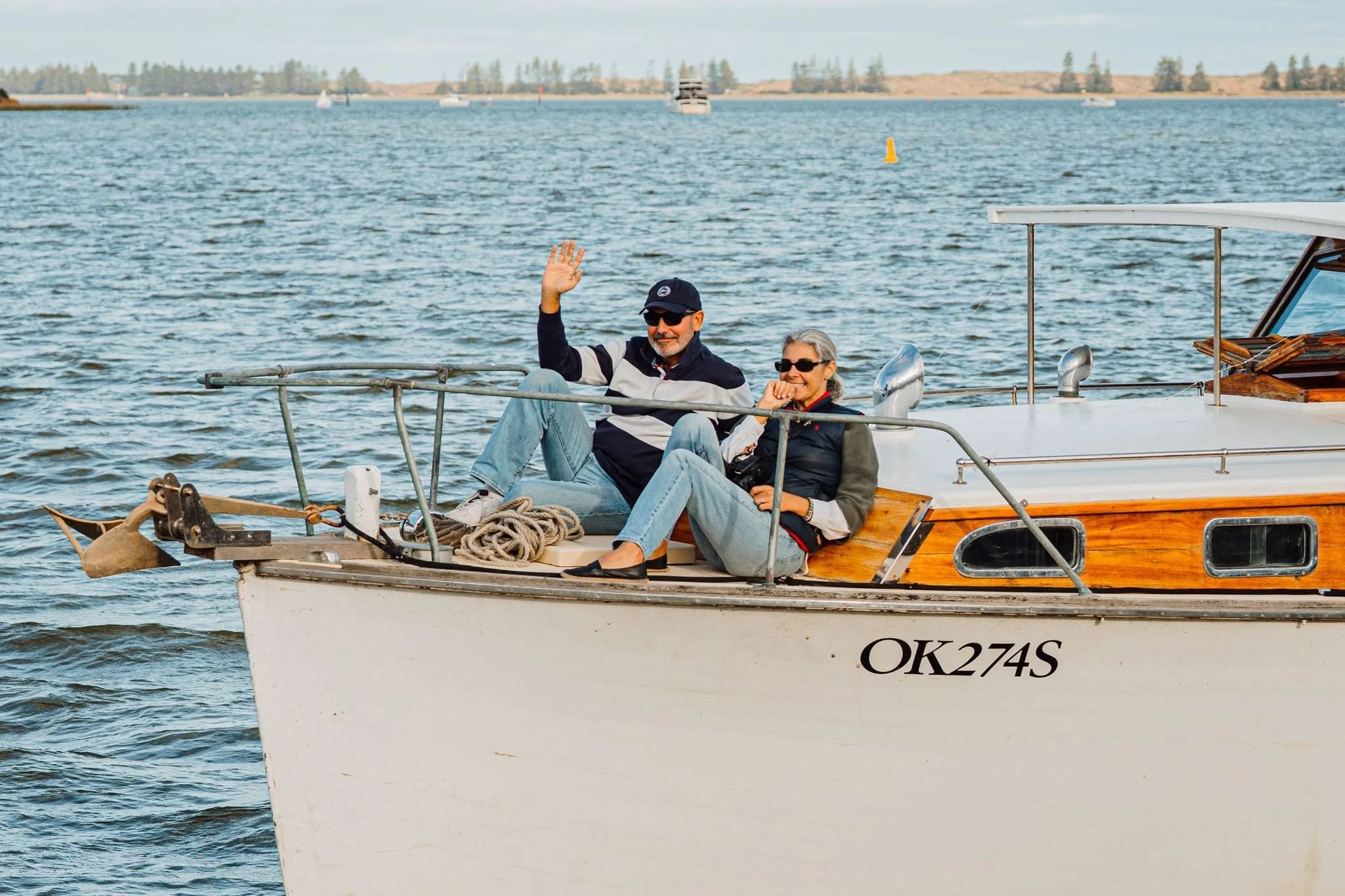 A man and woman sitting on the deck of a small boat, smiling and waving, with water and distant shoreline in the background.