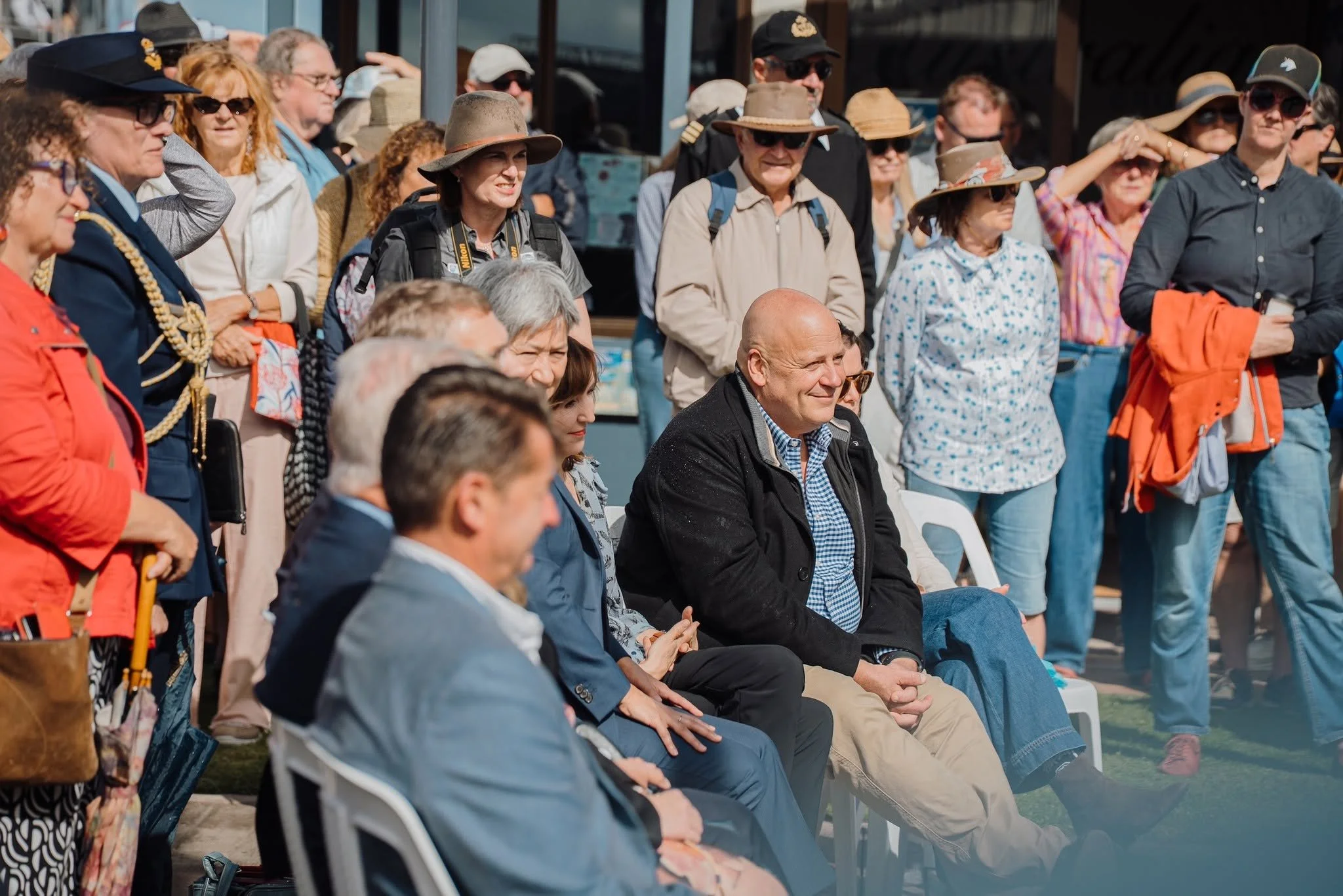 A group of diverse people sitting and standing outdoors, attending the Festival, with some wearing hats and sunglasses, under sunny weather.