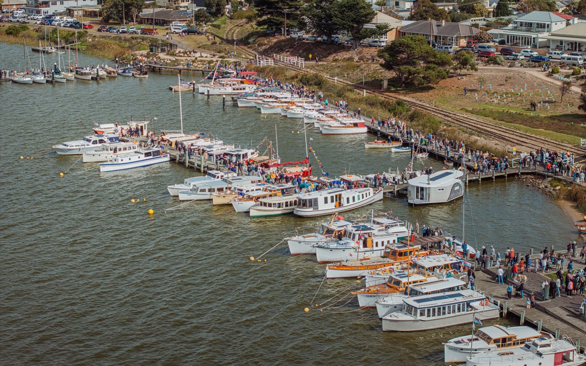 The busy Goolwa Wharf with numerous boats docked, many people walking on the dock, and a background of residential houses and trees near the shoreline.