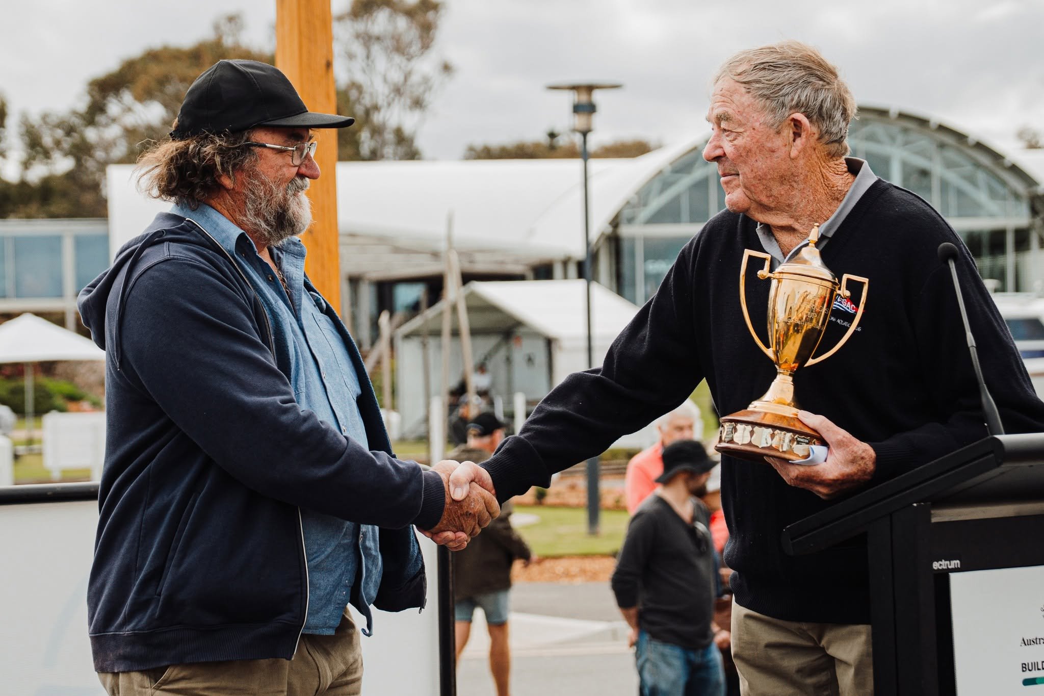 Two men shaking hands, one older man with gray hair holding a trophy, the other man wearing glasses and a black hat, at an outdoor event.