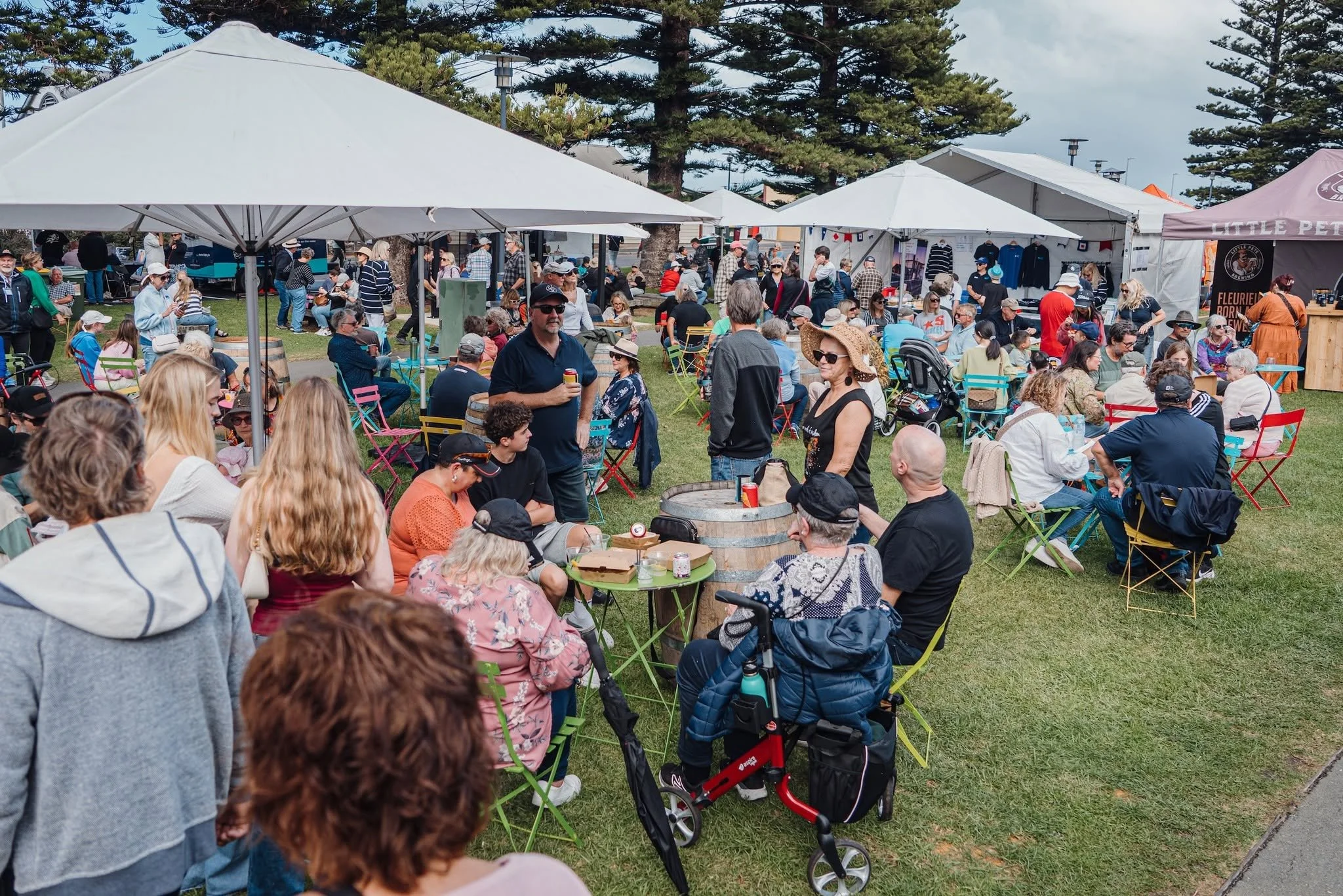 Crowd of people at the Festival with white tents, trees, and vendor booths in the background. Many are sitting on colourful chairs, some standing, and some walking around.
