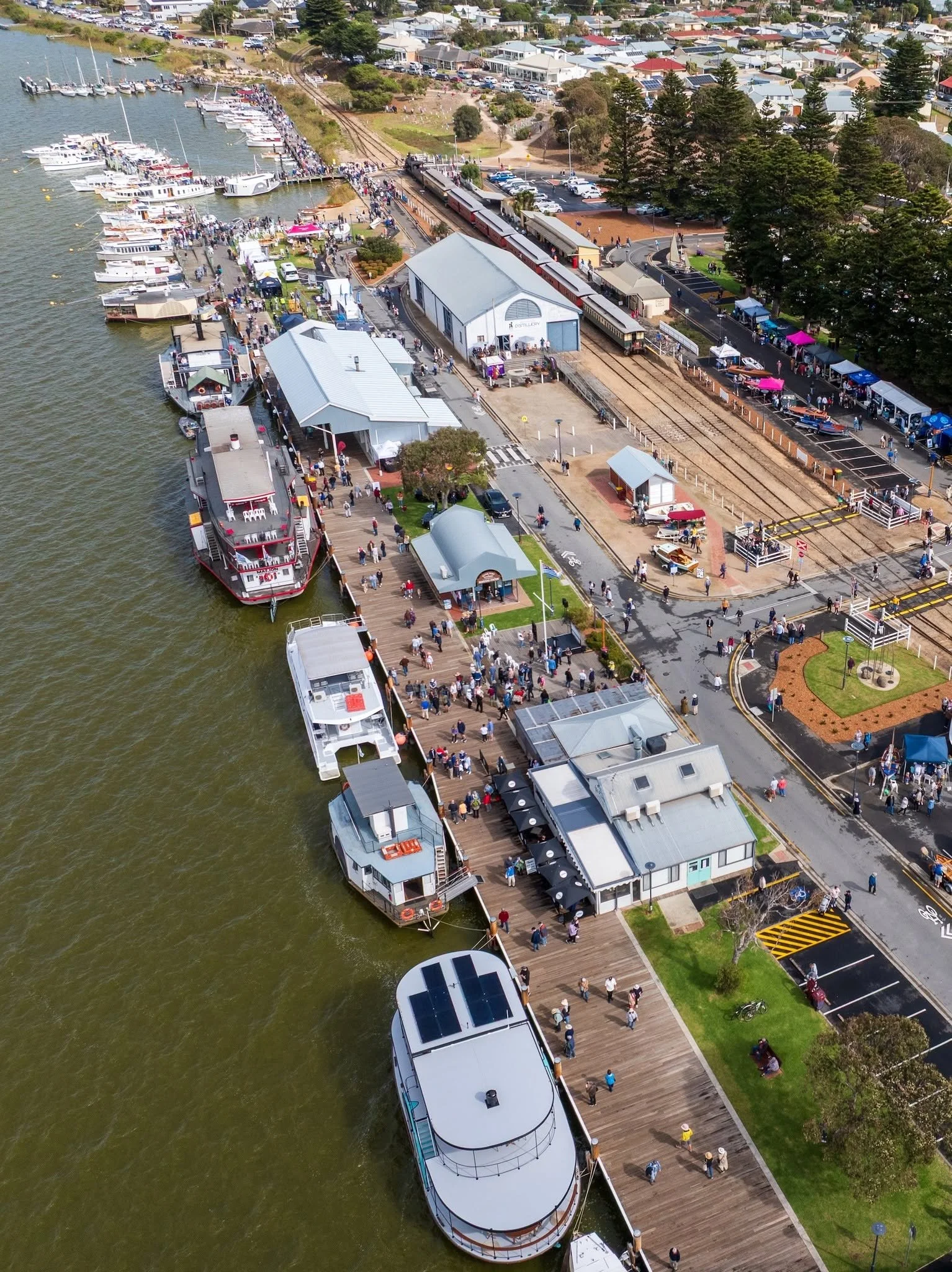 An aerial view of a waterfront area with boats docked, a busy market with tents and stalls, a steam train running through the area, and people walking along the boardwalk and nearby streets.