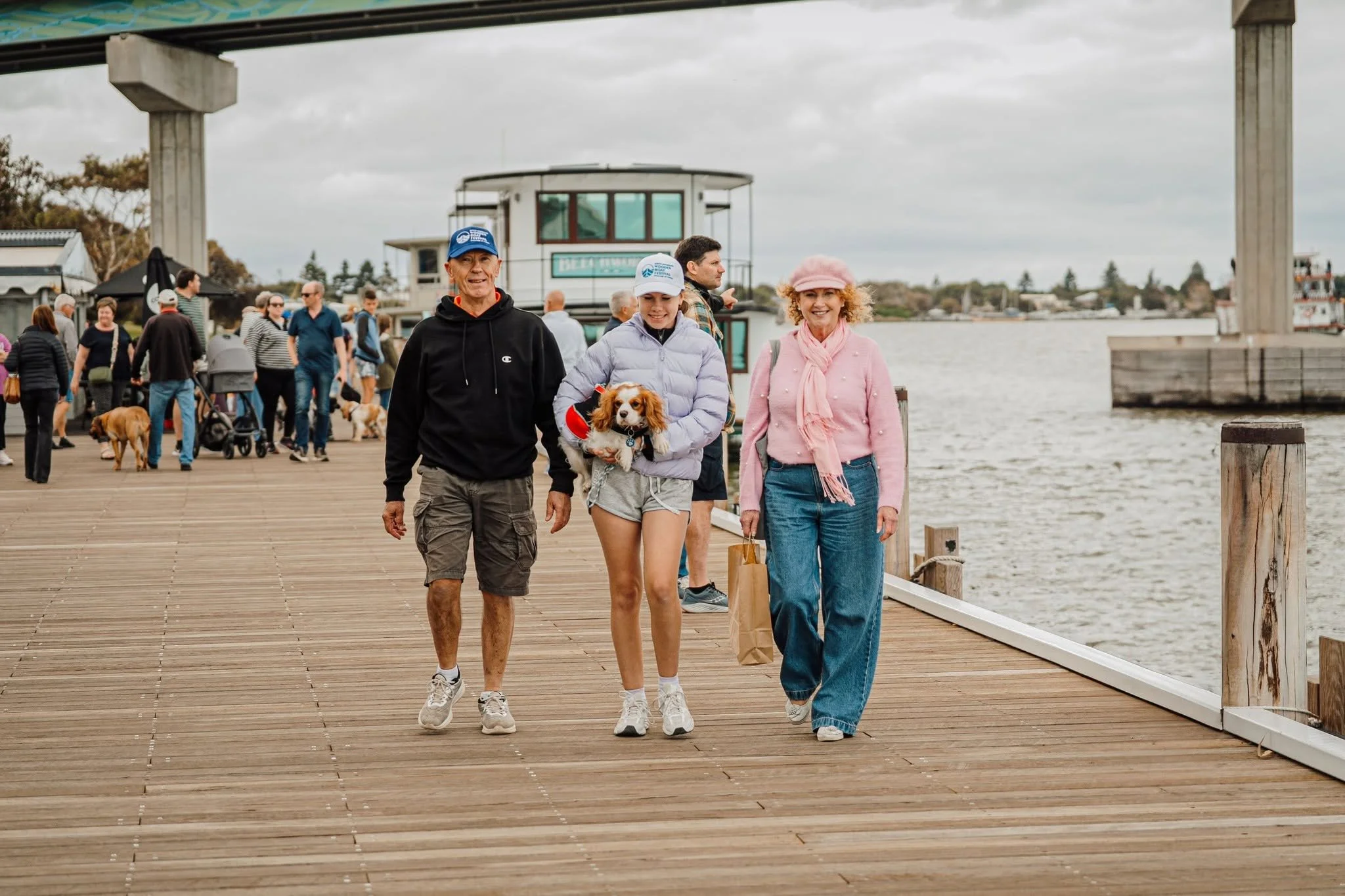 People walking on a wooden pier by the water, with a small building and other visitors in the background, as overcast skies fill the scene.