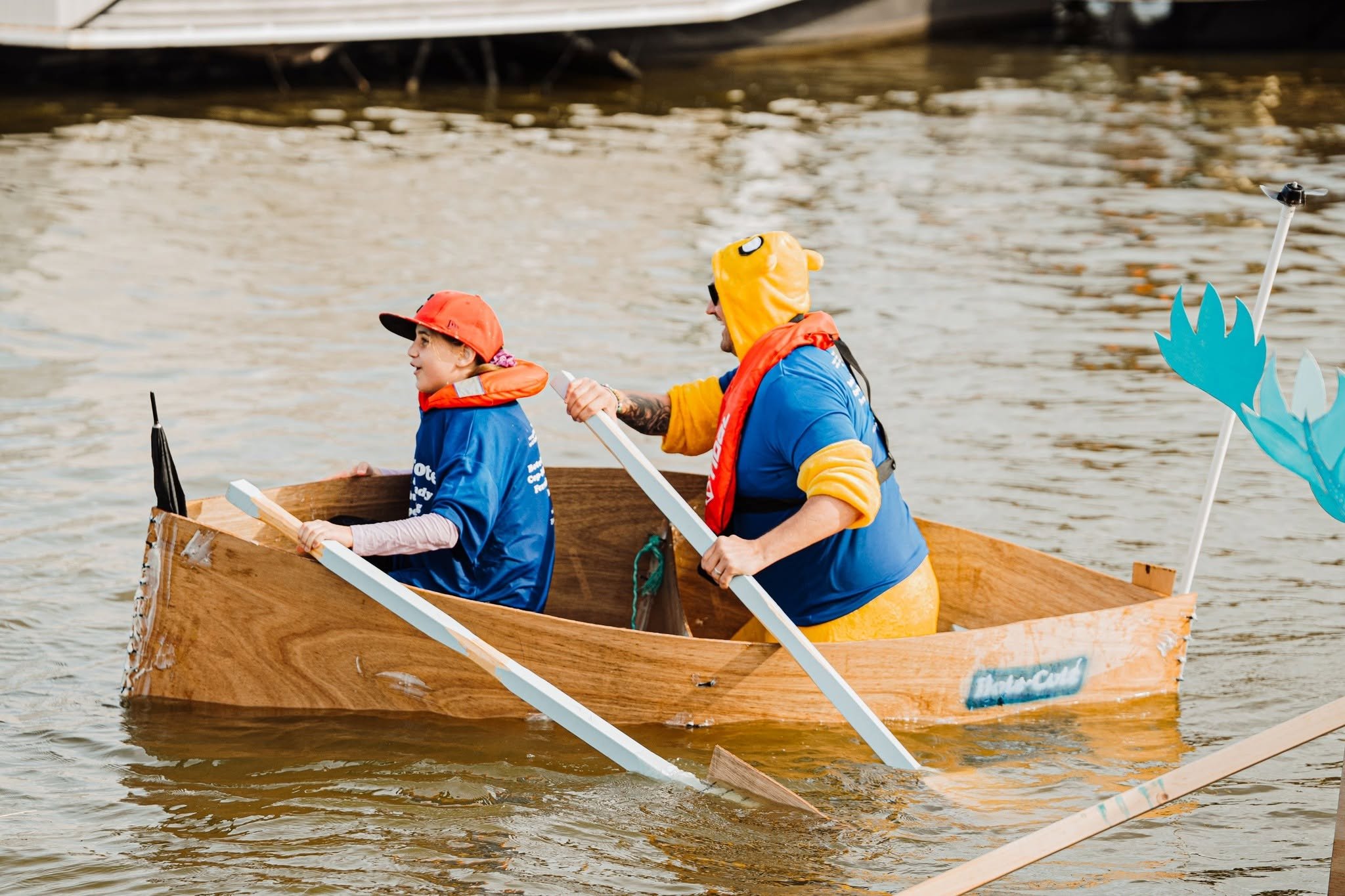 Two people in a wooden boat on a body of water, one wearing a yellow hoodie with animal ears and a life jacket, and the other wearing a red cap and blue shirt, holding paddles.