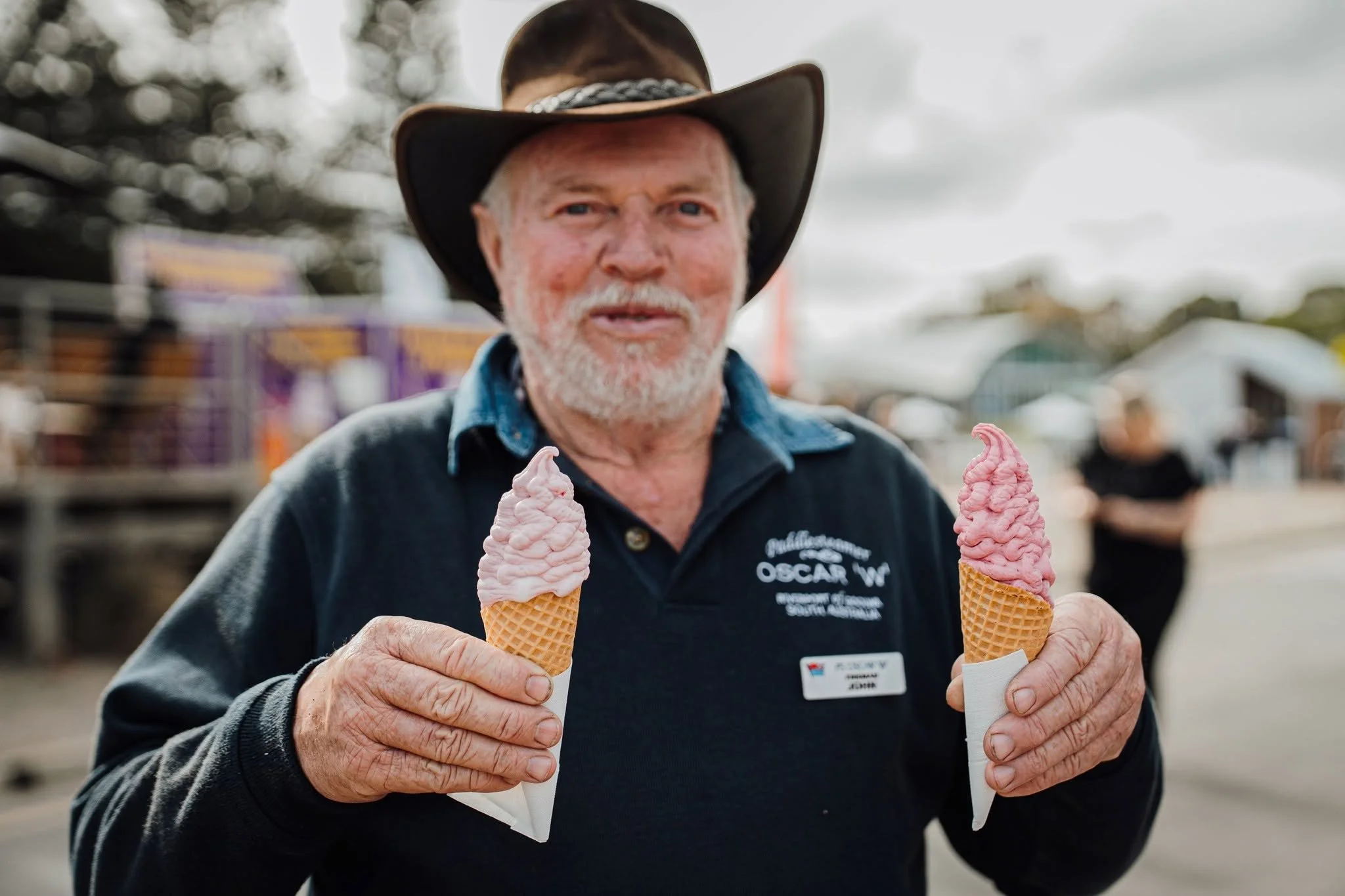 A man with a grey beard wearing a cowboy hat and a black jacket holding two pink-coloured ice cream cones at the Festival