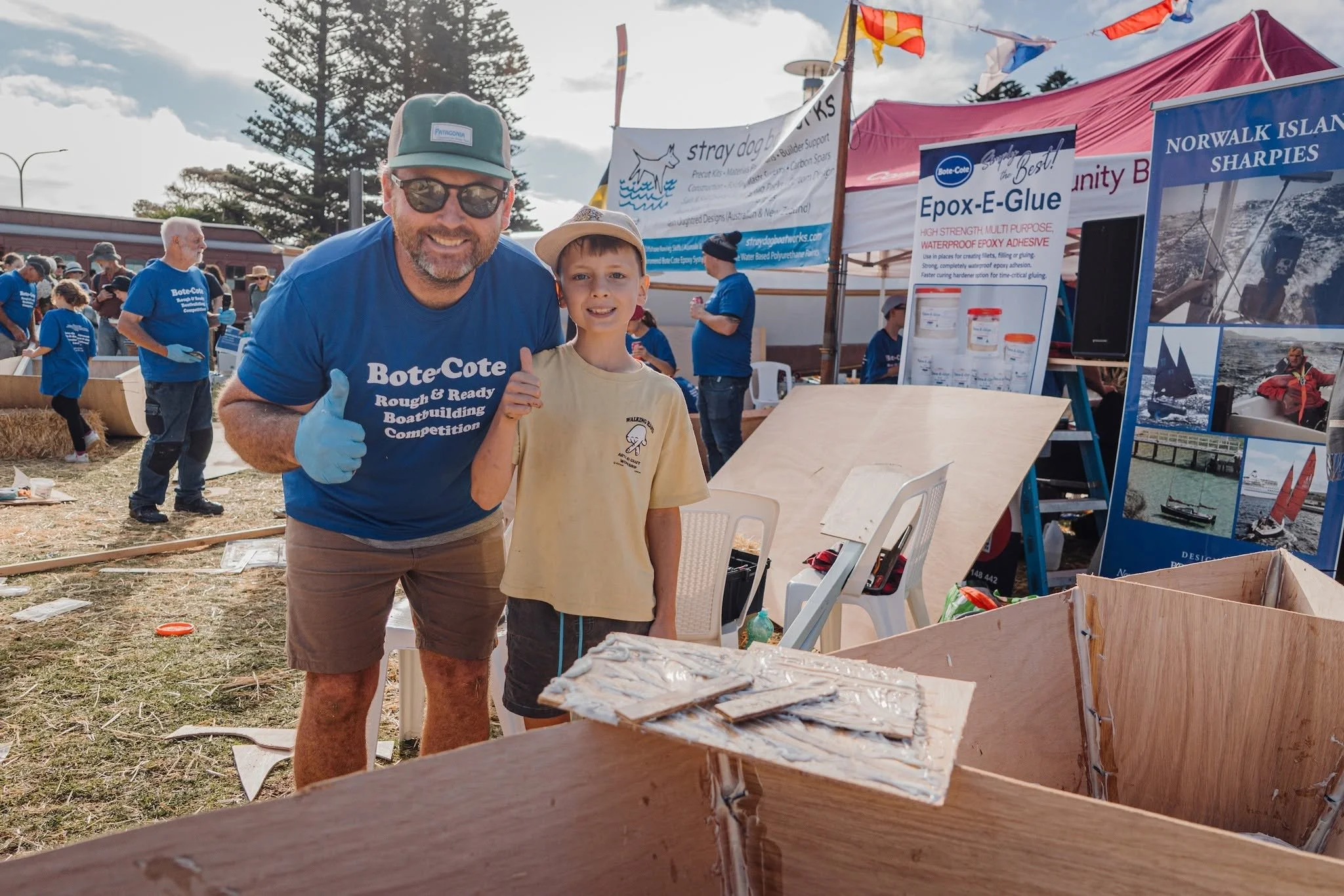 A man wearing sunglasses, a green cap, and a blue t-shirt is smiling and giving a thumbs-up next to a young boy in a tan t-shirt and a wide-brimmed hat. They are at the Rough'n'Ready boat building event.